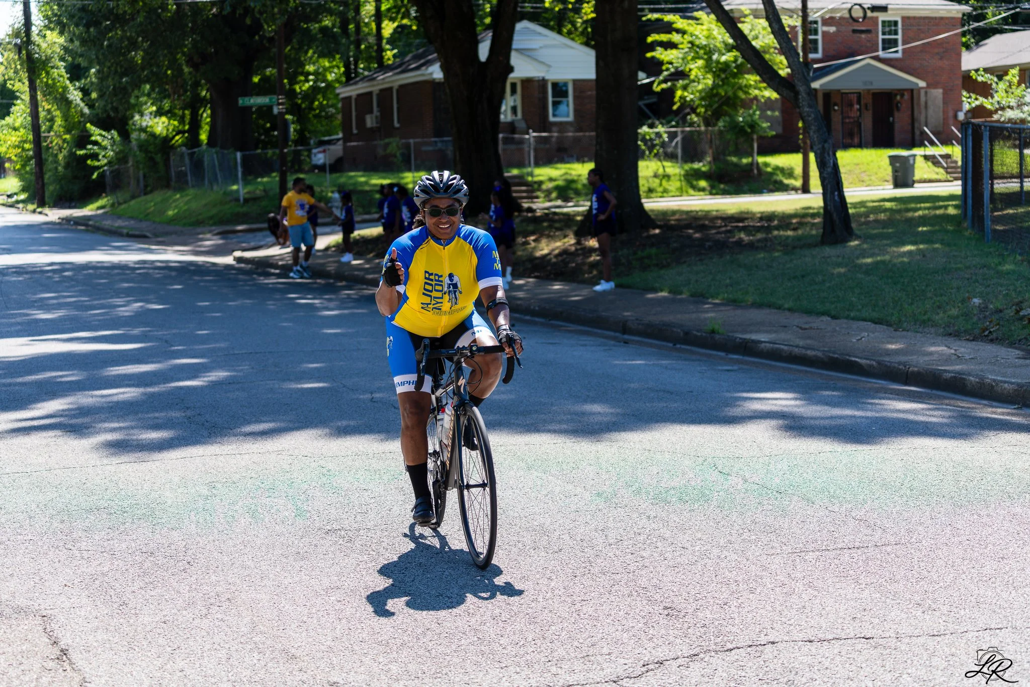 A woman in cycling gear riding a bicycle on the street, smiling and giving a thumbs up. In the background, children are gathered under trees on a sunny day in a residential neighborhood.