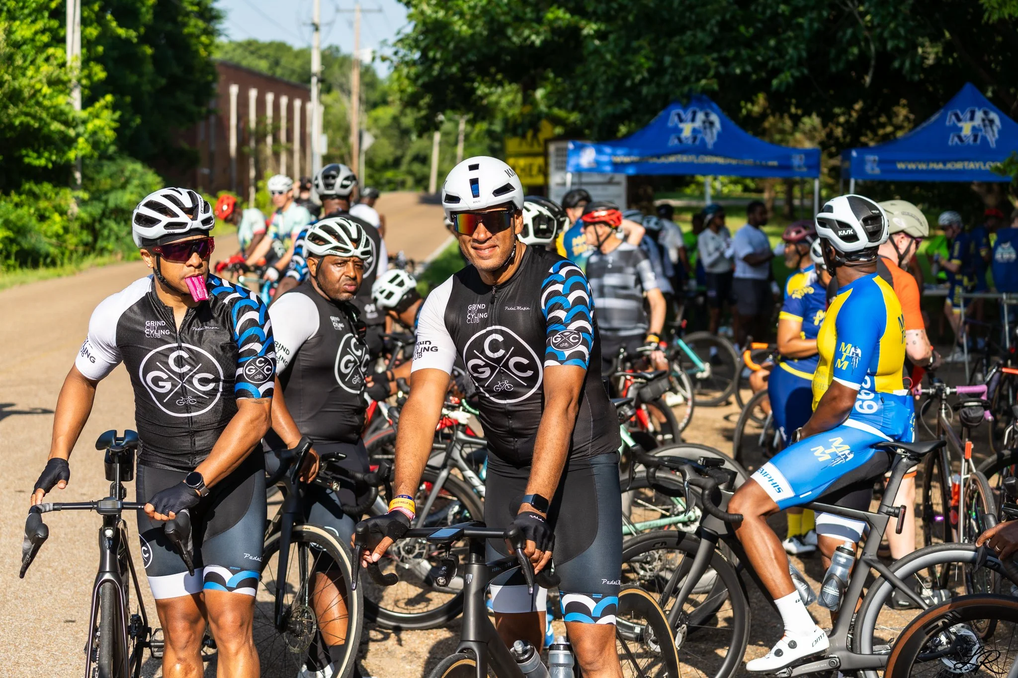 Group of cyclists at a cycling event, wearing helmets and cycling gear, standing outdoors with tents and trees in the background.
