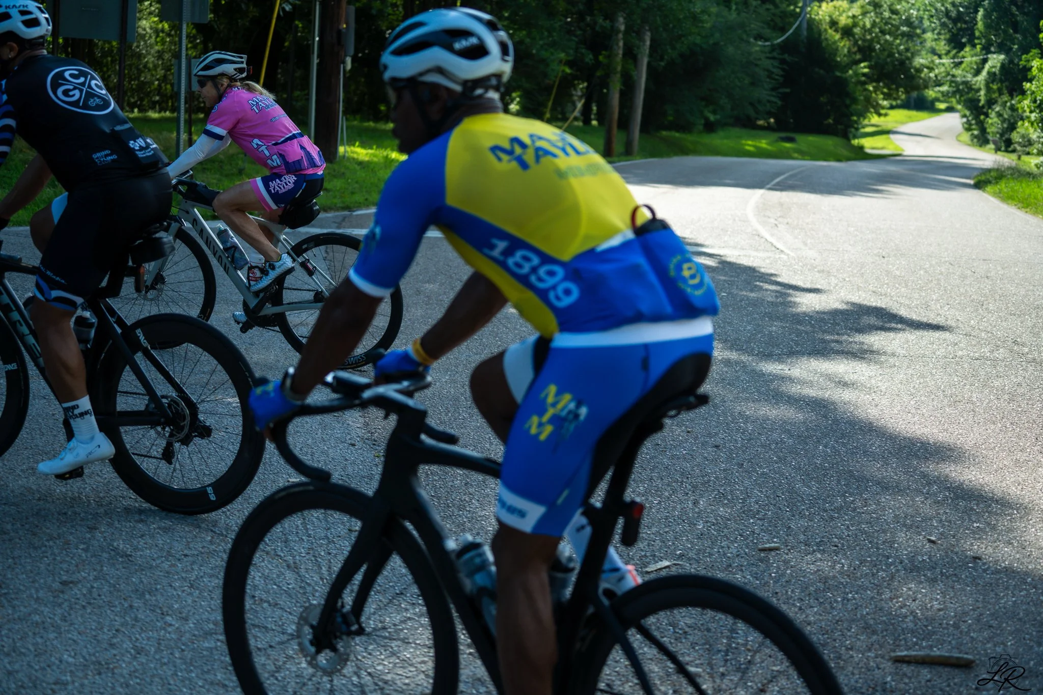 Group of cyclists riding on a winding road surrounded by greenery, wearing helmets and colorful cycling jerseys, in a sunny outdoor setting.