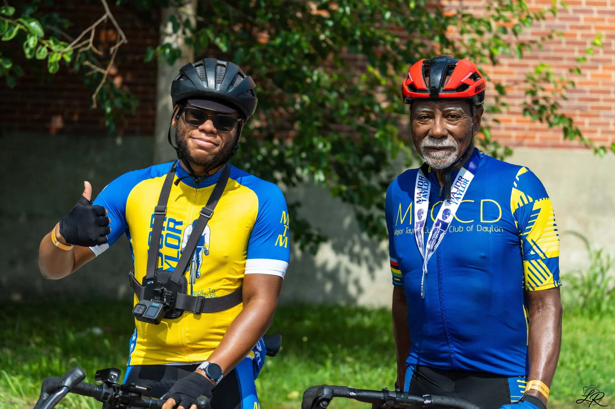 Two men wearing cycling gear and helmets standing outdoors next to their bicycles, smiling and giving a thumbs-up. One man is giving a thumbs-up with his thumb, while the other is standing on a grassy area with trees and a brick wall background.