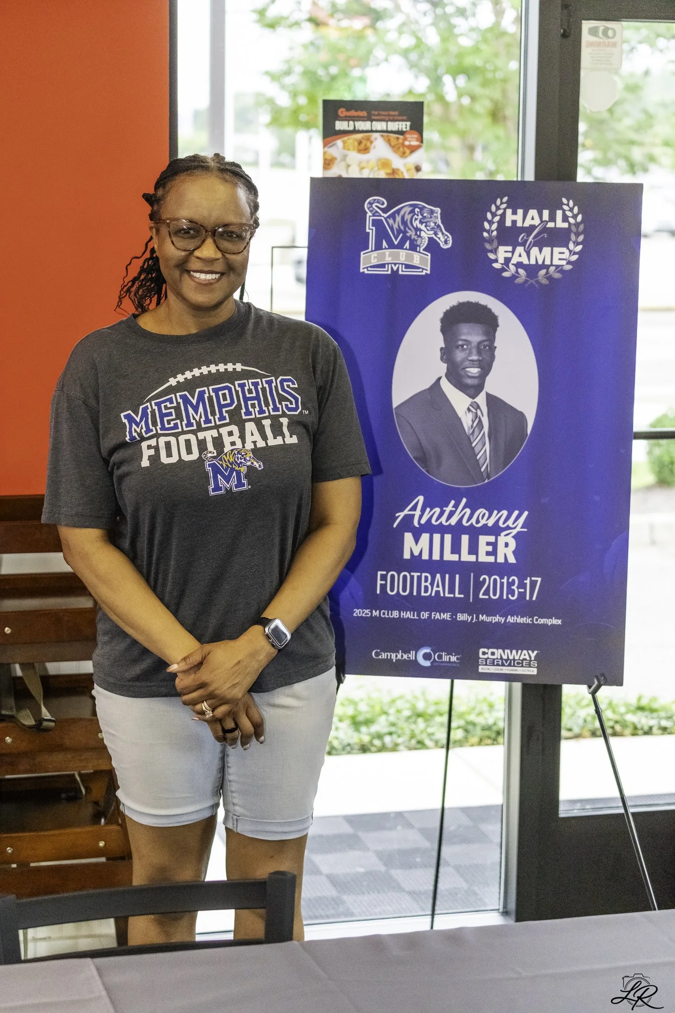 A woman smiling and standing next to a blue Hall of Fame poster for Anthony Miller, football player from 2013-2017, at an indoor event.