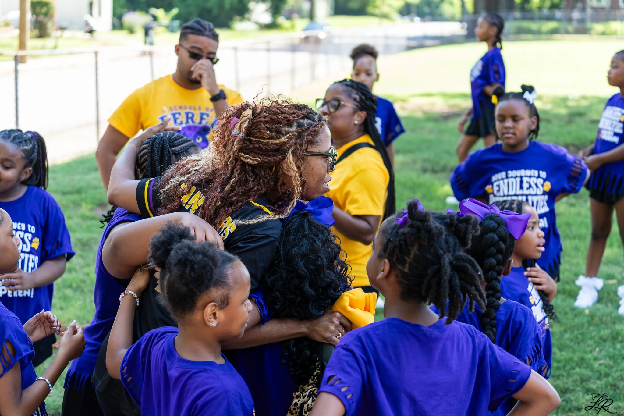 Group of children and adults with dark skin participating in an outdoor event, wearing blue and yellow shirts, some embracing and interacting with each other on a grassy area with trees and a fence in the background.