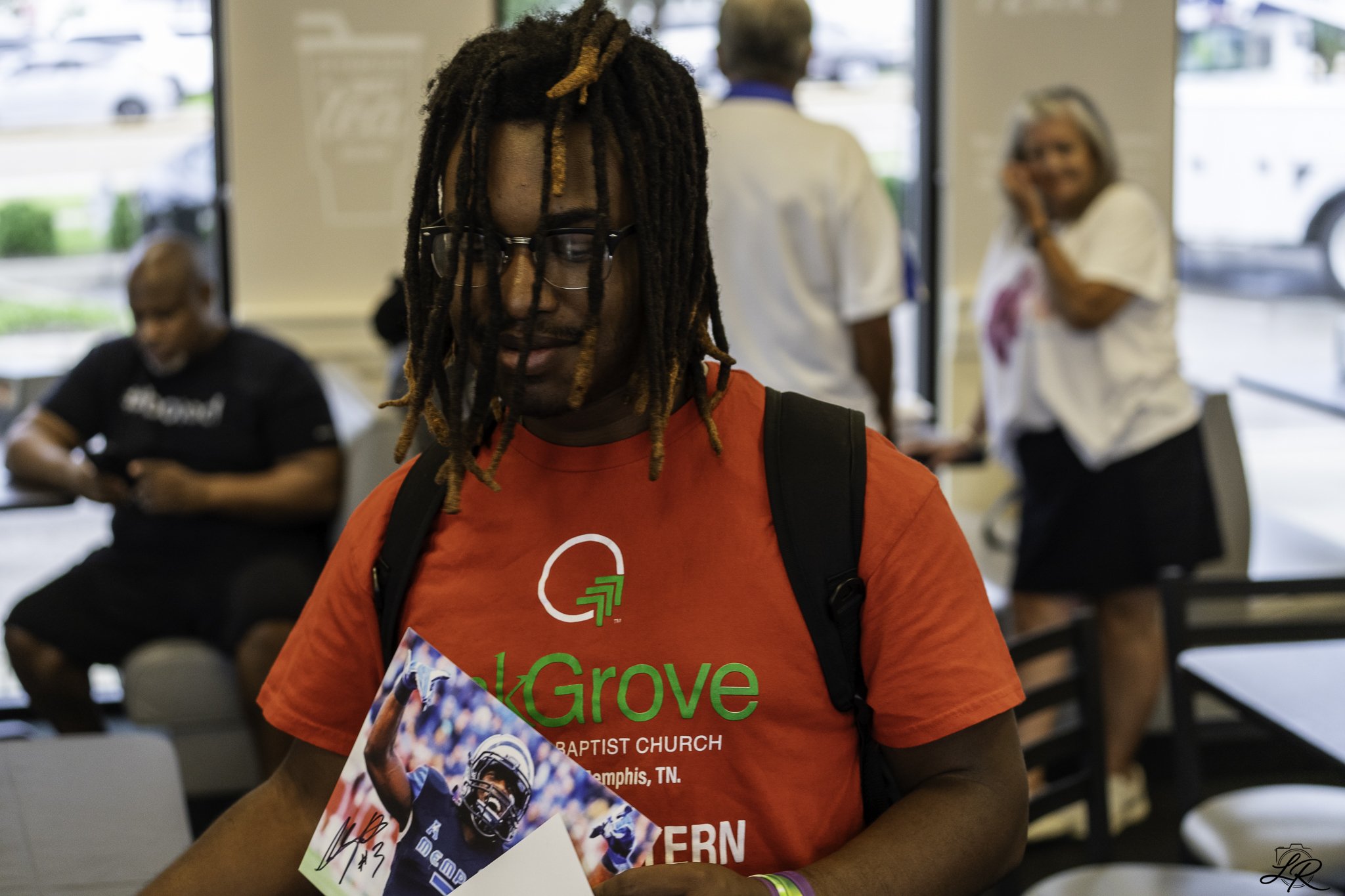 A person with long dreadlocks and glasses wearing a red t-shirt with text, holding a pamphlet and a signed football, standing inside a building with people in the background.