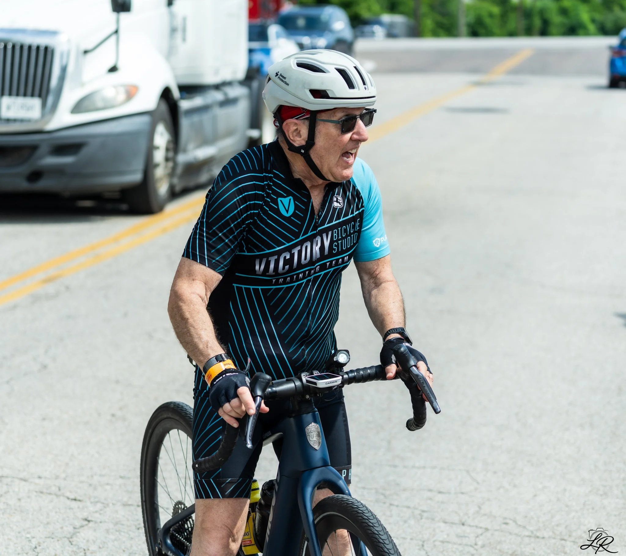 A man riding a bicycle wearing a white helmet and sunglasses, wearing a black and blue cycling jersey, on a street with parked cars and green trees in the background.