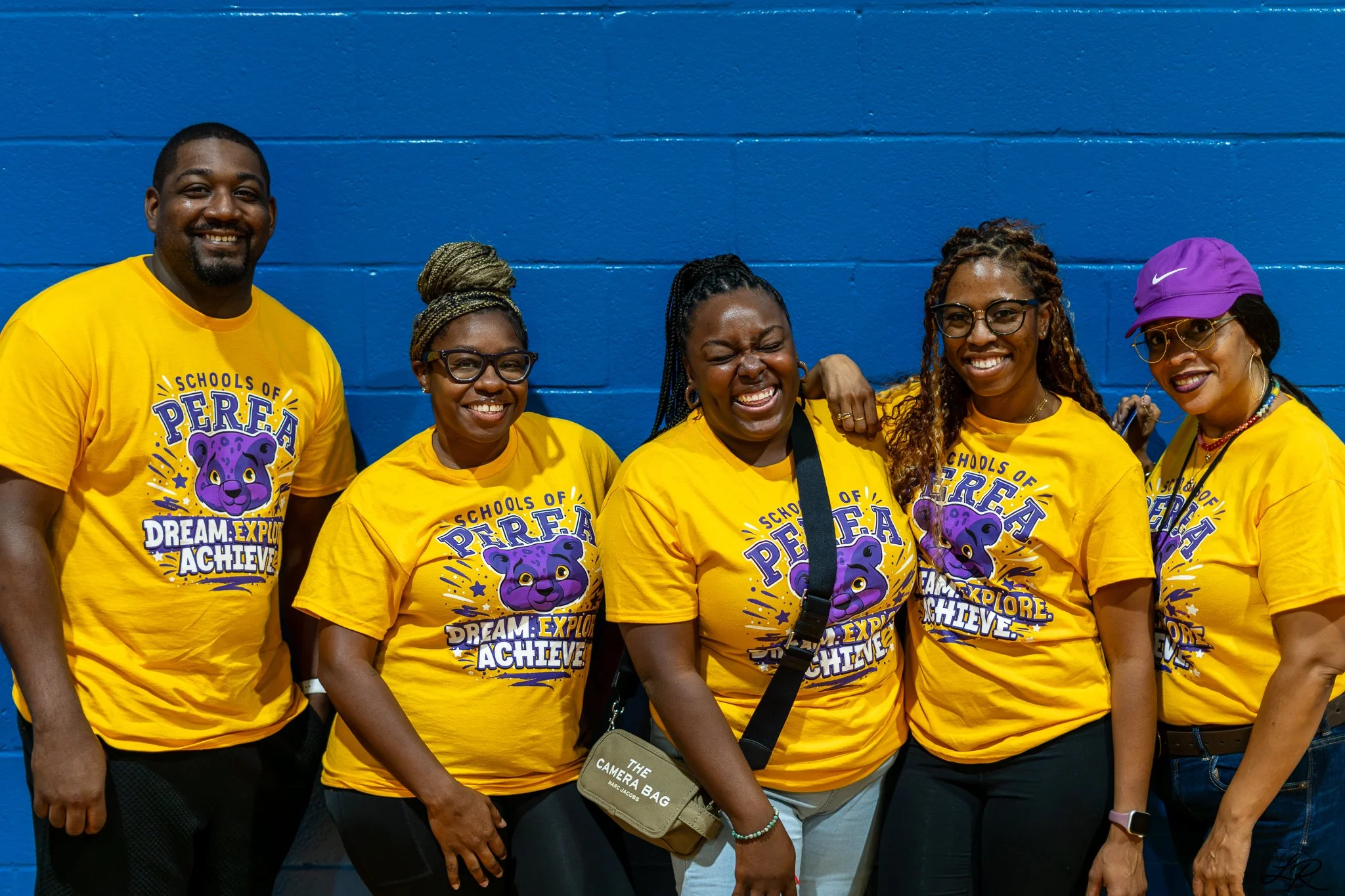 Group of six smiling people wearing yellow T-shirts with a purple panther graphic and text, standing against a blue wall.