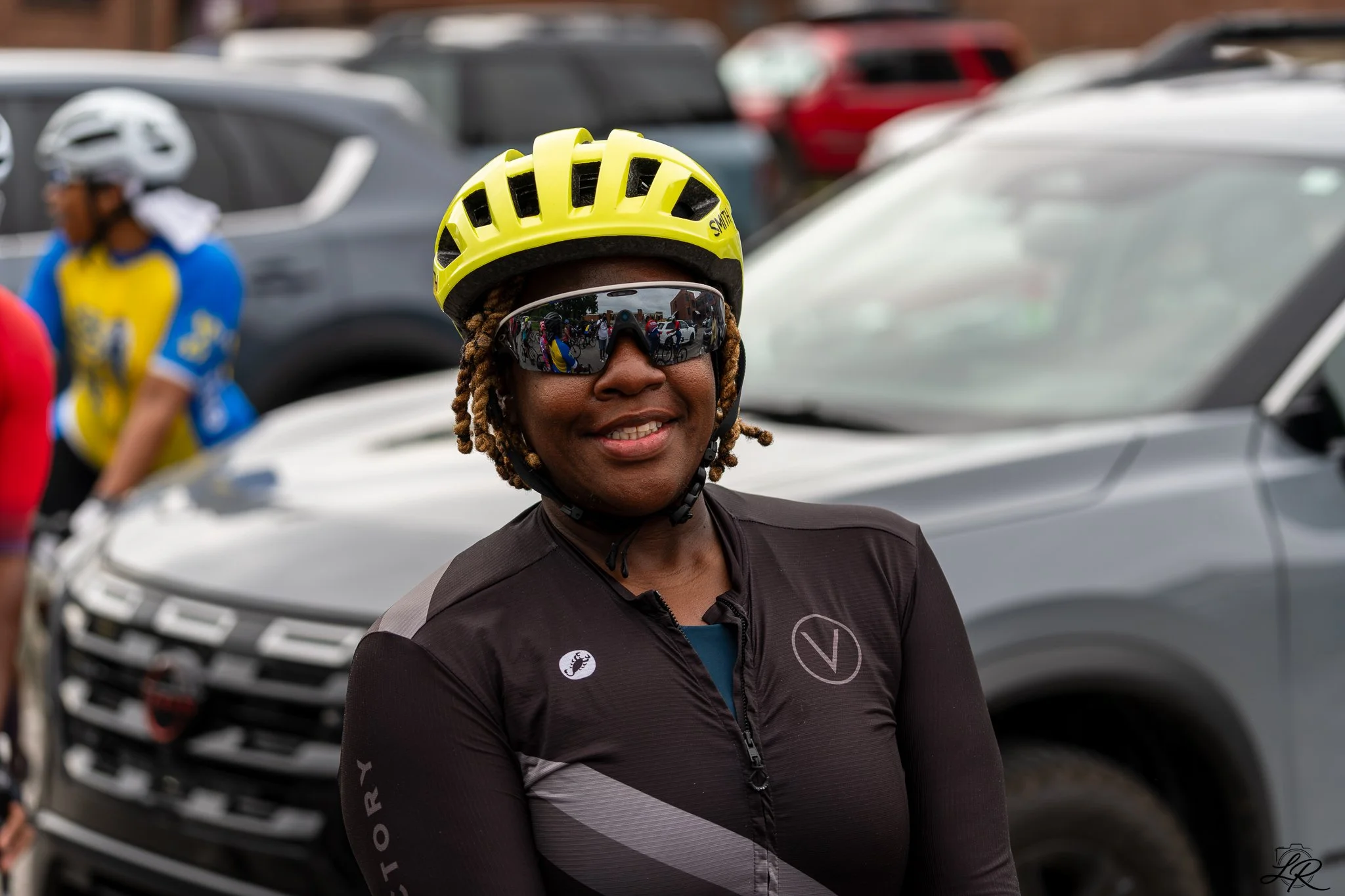 A woman in cycling gear wearing a yellow helmet and reflective sunglasses, smiling, with several parked cars in the background.