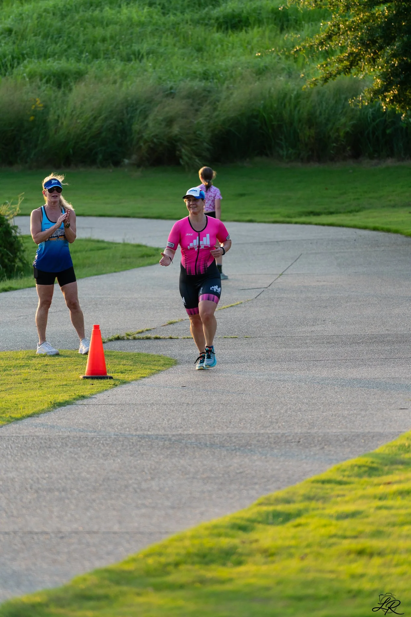 A woman running outdoors near a grassy area with two women clapping and watching her. There is a traffic cone on the ground, and another woman is walking in the background. The sun is setting, casting warm light.