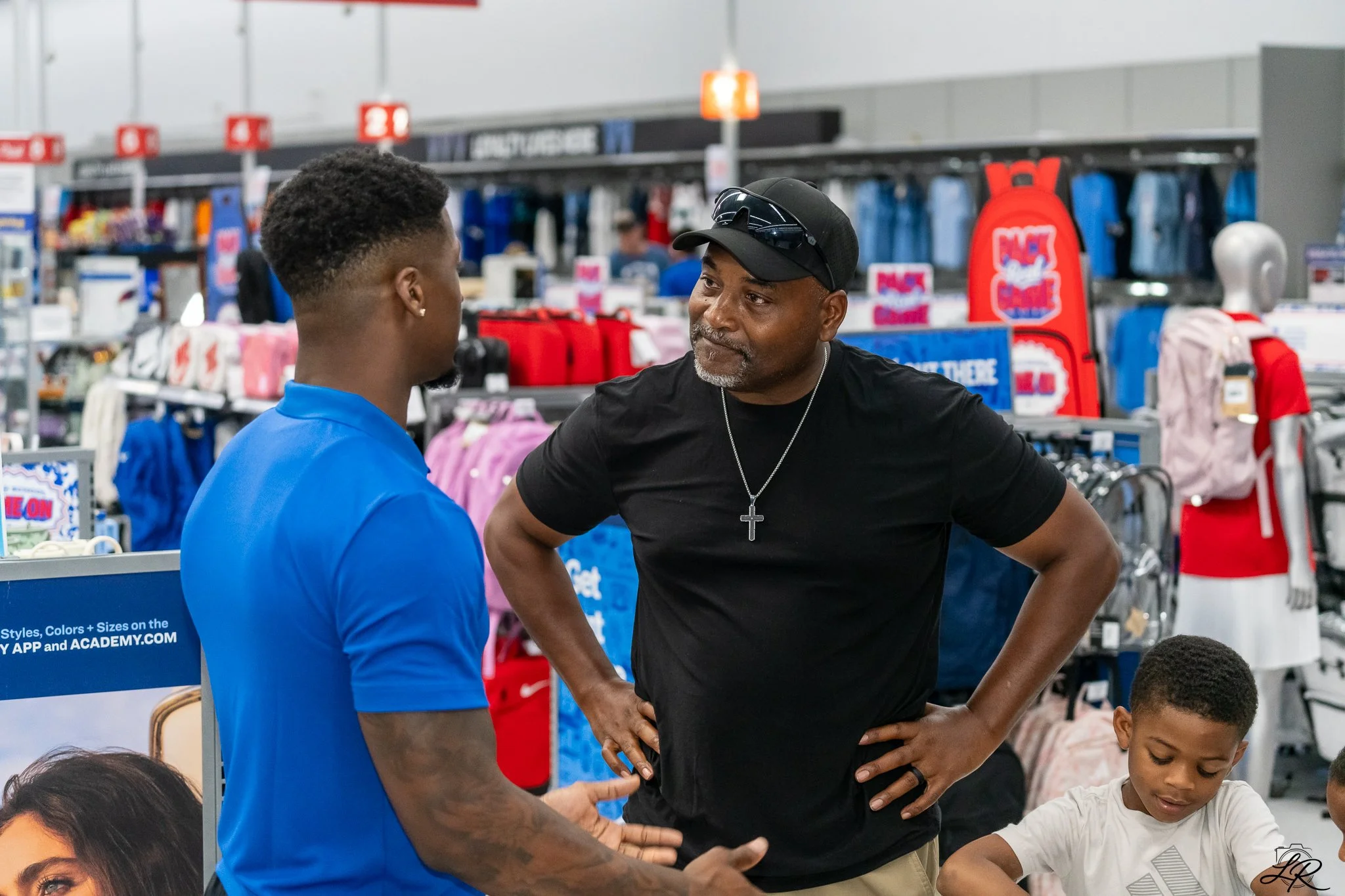 Two men engaged in a conversation inside a retail store, with colorful backpacks and merchandise in the background.