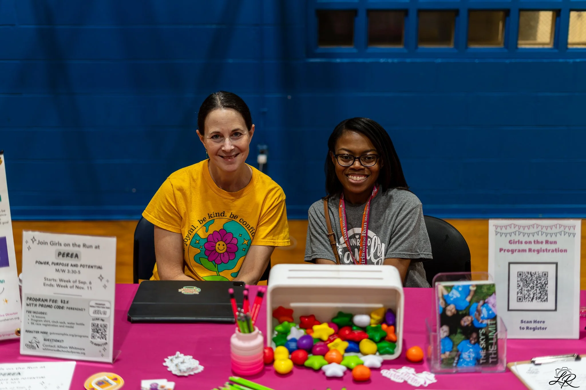 Two women sitting behind a registration table at an event, smiling. The woman on the left is wearing a yellow T-shirt with a colorful flower design, and the woman on the right is wearing glasses and a gray T-shirt. The table has various colorful game