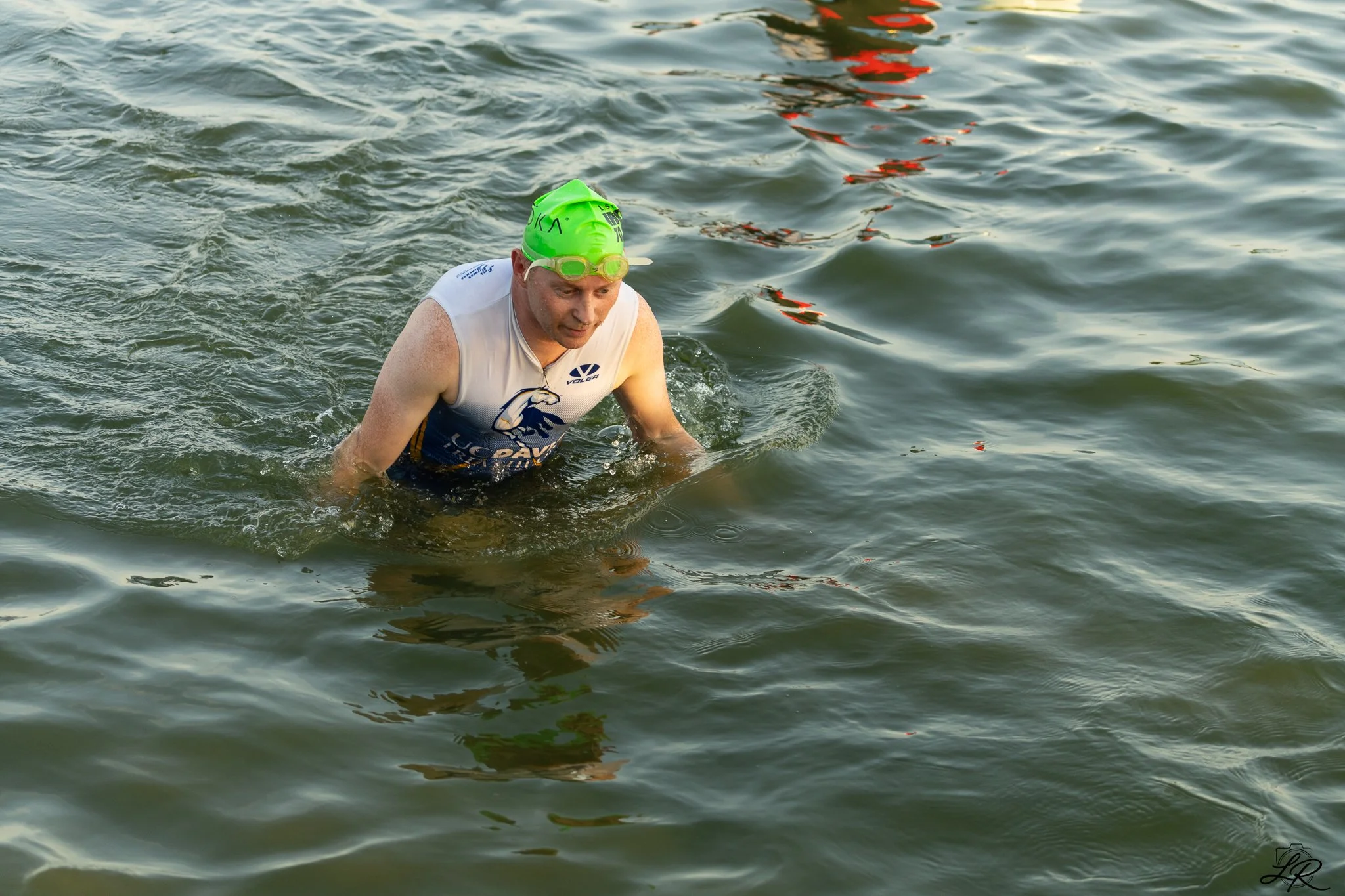 A man wearing a green swim cap and goggles is emerging from the water, likely after a swim or triathlon, with the water surrounding him. He is wearing a white athletic top with logos.