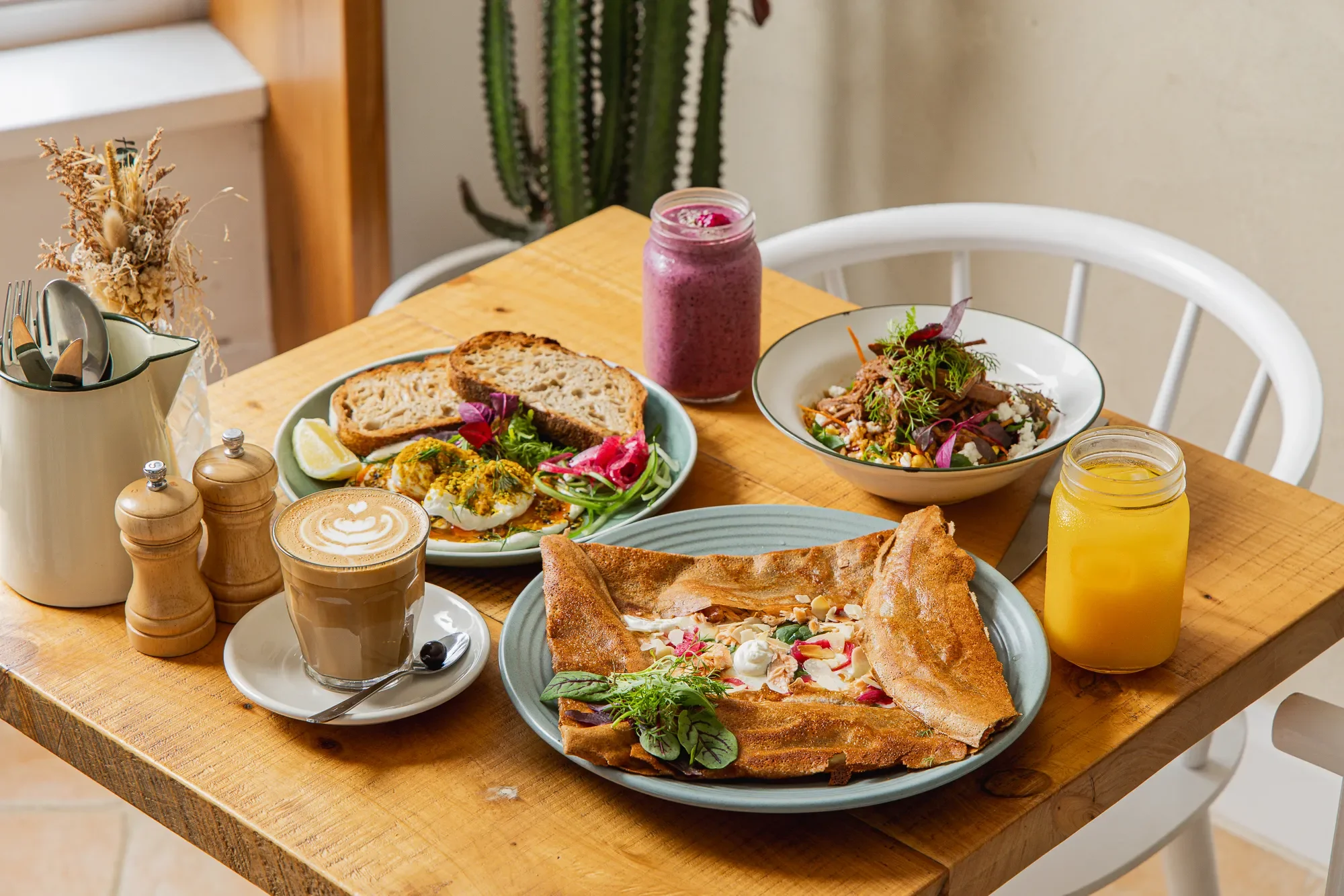 A wooden table set with various breakfast foods including a slice of toast, a salad with greens and vegetables, a crepe filled with fruit and cream, a baked item, and two beverages, one of an iced berry smoothie and the other of a latte with foam art