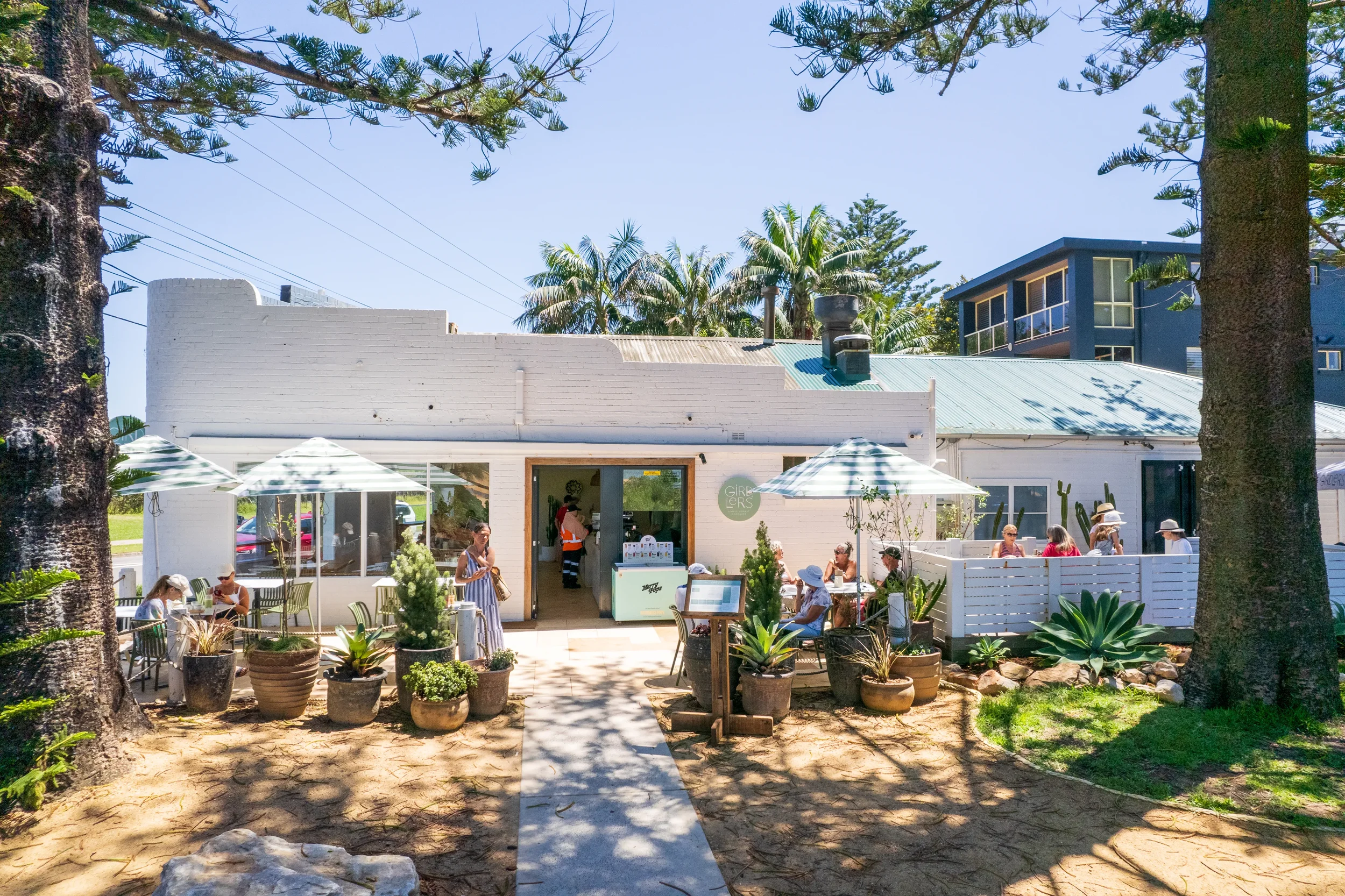 Outdoor cafe scene with people sitting at tables, greenery, and trees in front of a white building with an adjacent modern blue building, under a clear blue sky.