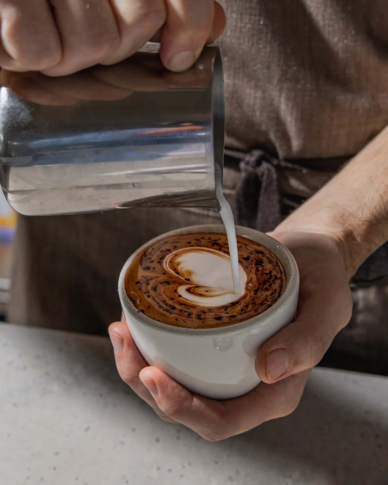 A person holding a white cup filled with a mocha coffee topped with whipped cream and chocolate shavings, while pouring steamed milk into the coffee.