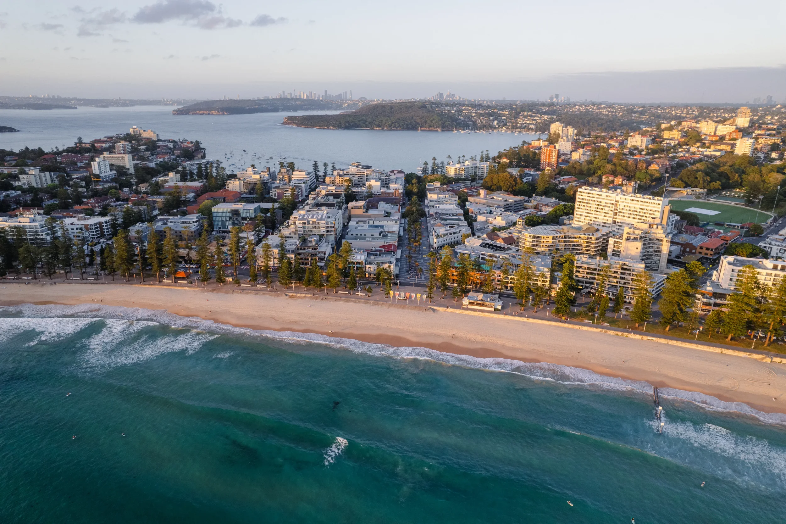 Aerial view of a city coastline with sandy beach in the foreground, high-rise buildings, a bay with boats, and green hilly land in the background.