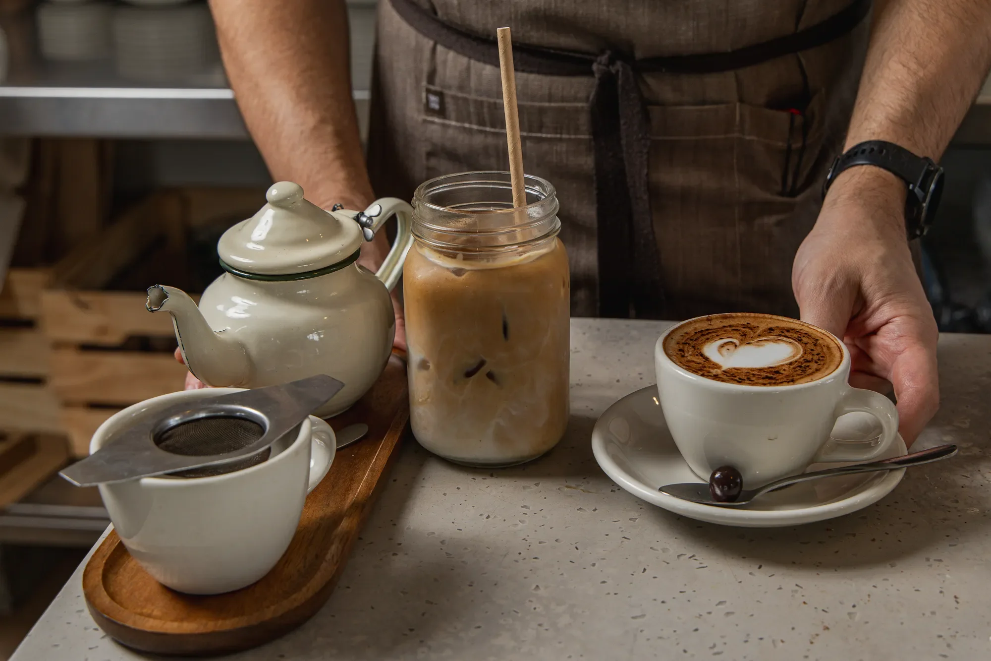 A person wearing a brown apron preparing coffee drinks on a kitchen countertop. There are two cups of cappuccino with latte art, a jar of iced coffee with a straw, and a beige teapot on a wooden tray with a metal strainer at the front.