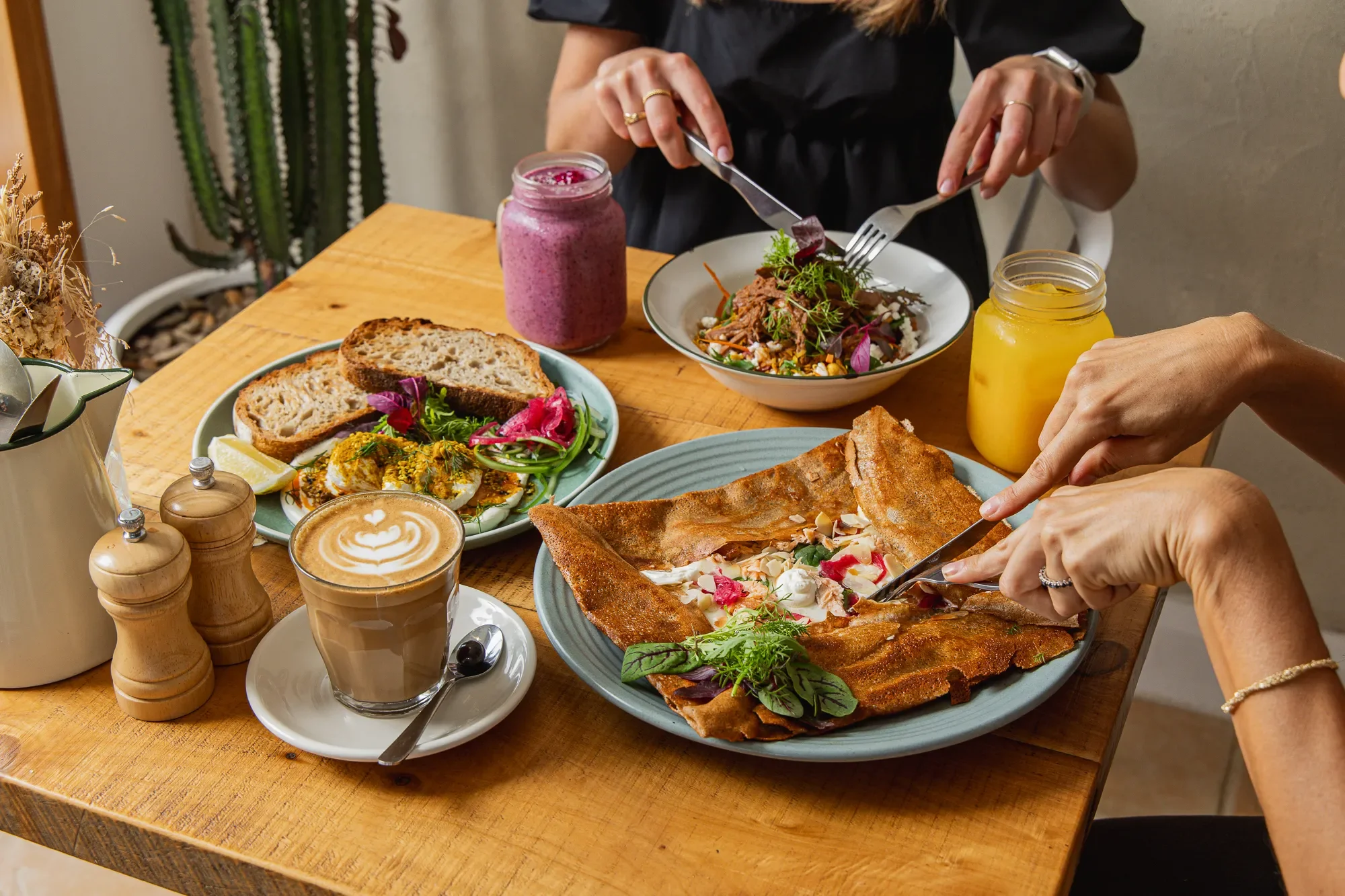 A table with various foods including a pizza, salad, bread, a pink smoothie, a yellow drink, a latte, and condiments, with two people serving themselves at the table.