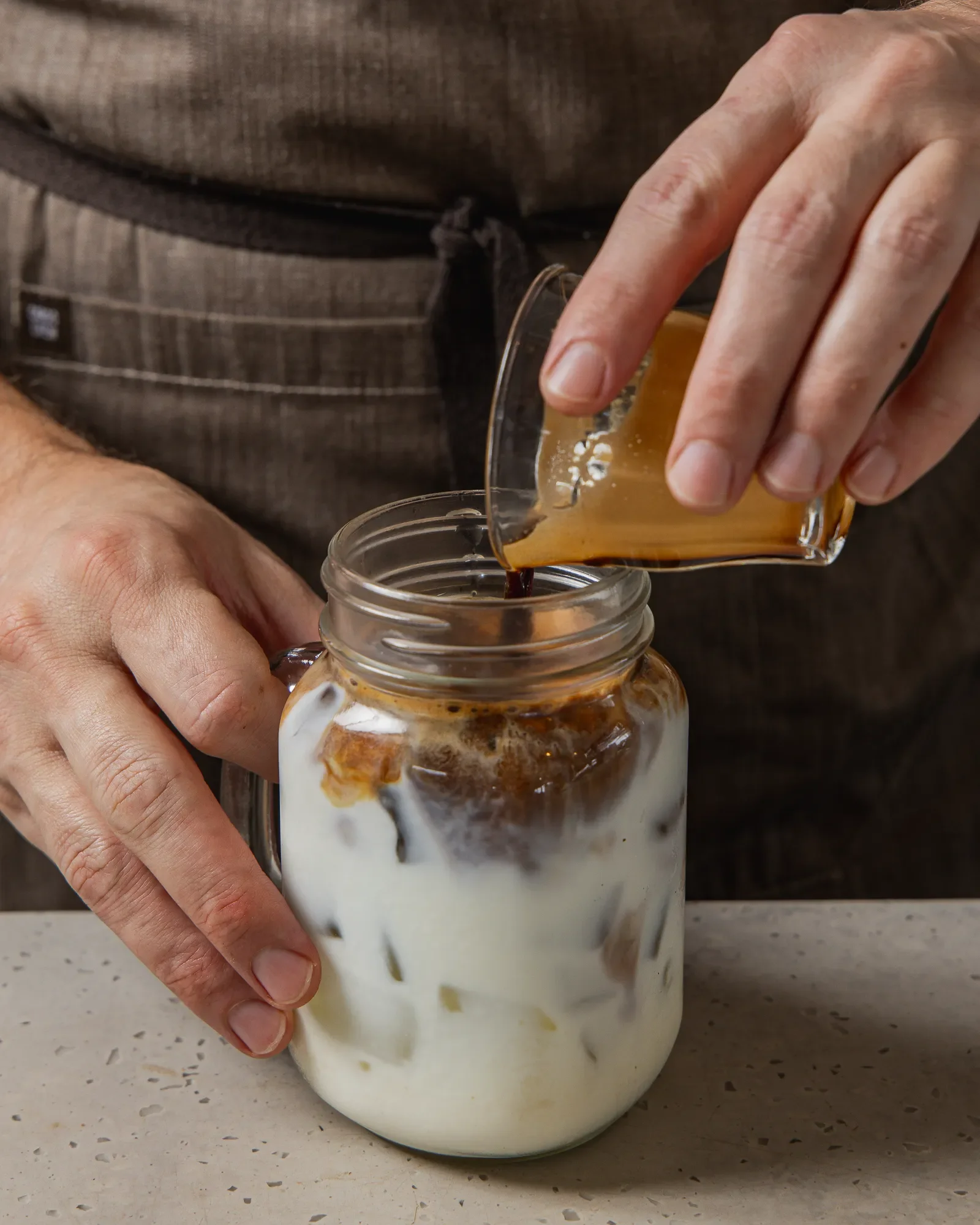 Person pouring coffee into a glass jar with milk at the bottom, cream and coffee swirls visible.