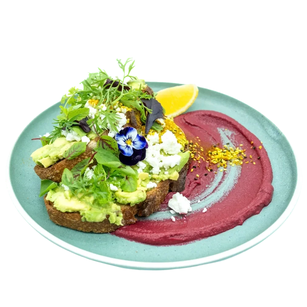 A plate with toast topped with avocado, microgreens, edible flowers, and crumbled cheese, accompanied by a lemon wedge and a smear of beetroot puree garnished with crushed nuts.