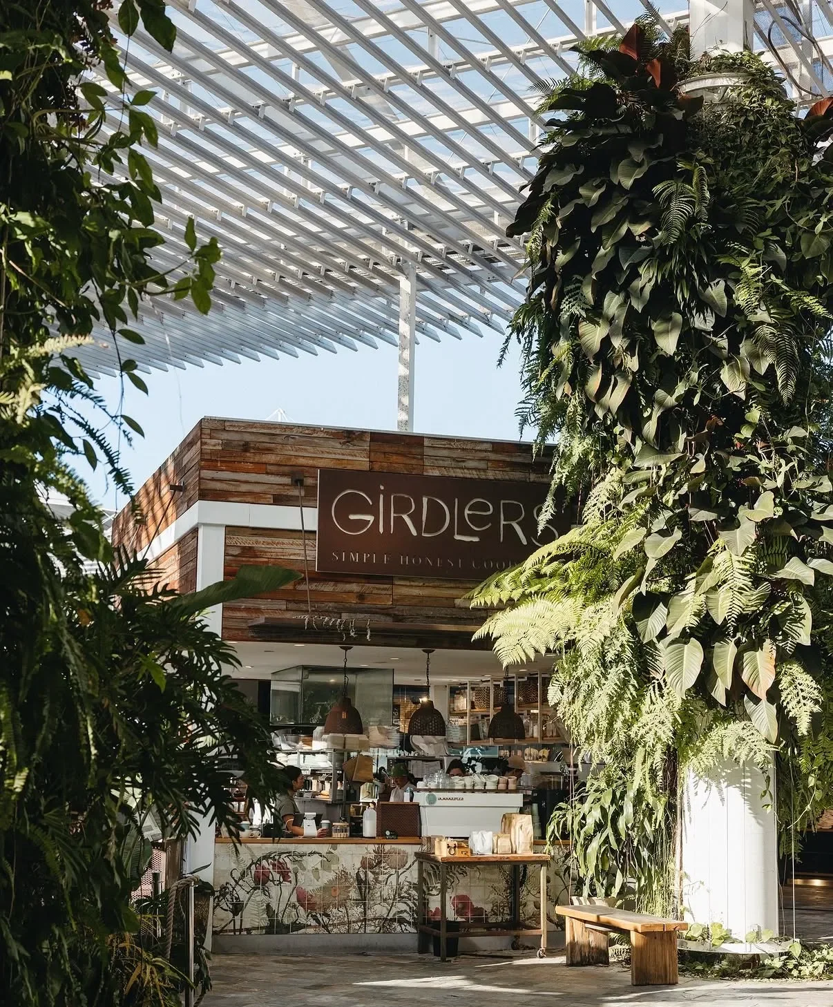 View of a cafe called GIRDLE with a modern wooden facade, lush greenery in the foreground, and a glass roof overhead letting in sunlight.