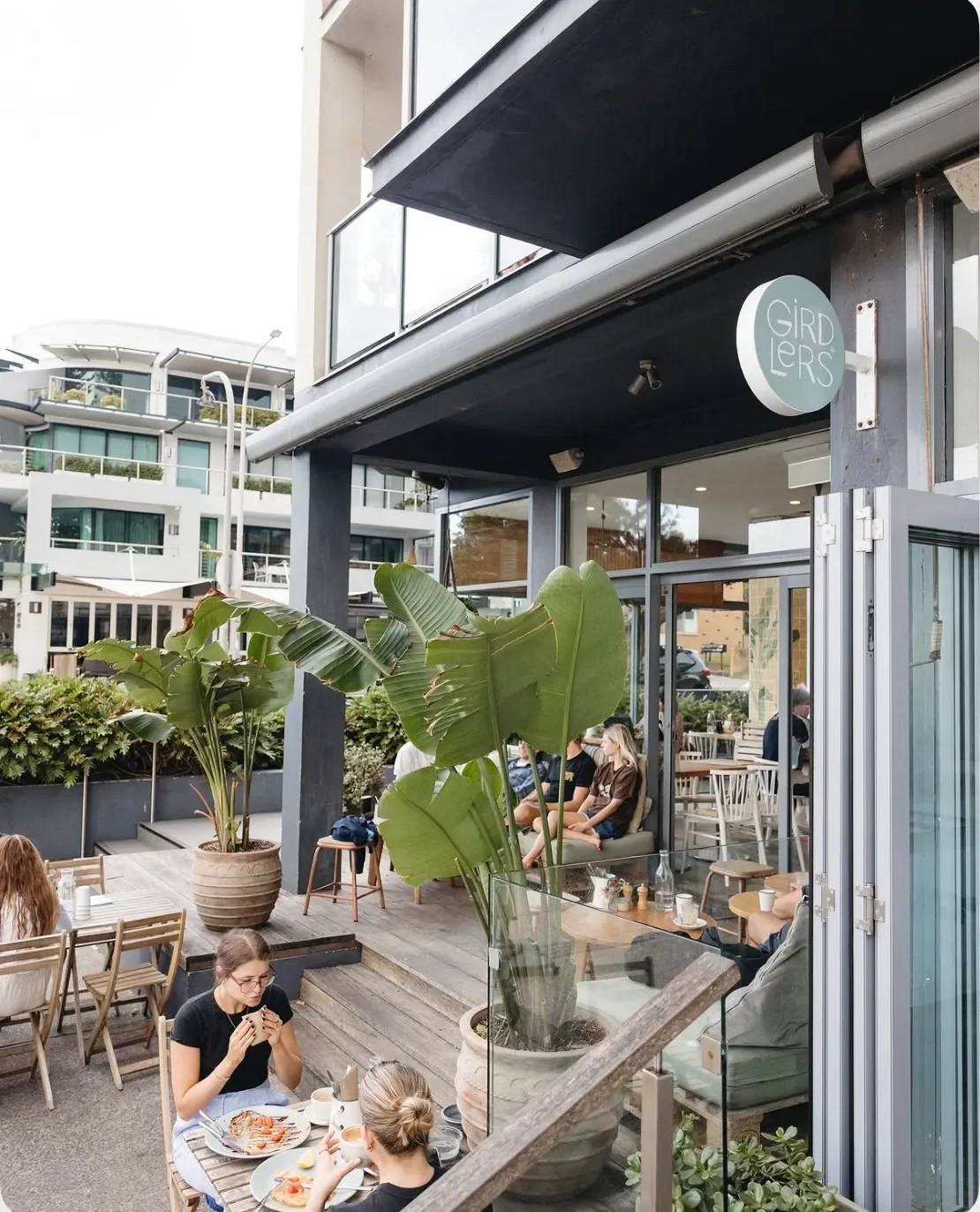Outdoor seating area of a modern cafe with people dining, large potted plants, and a sign that says 'Gird Glers'.