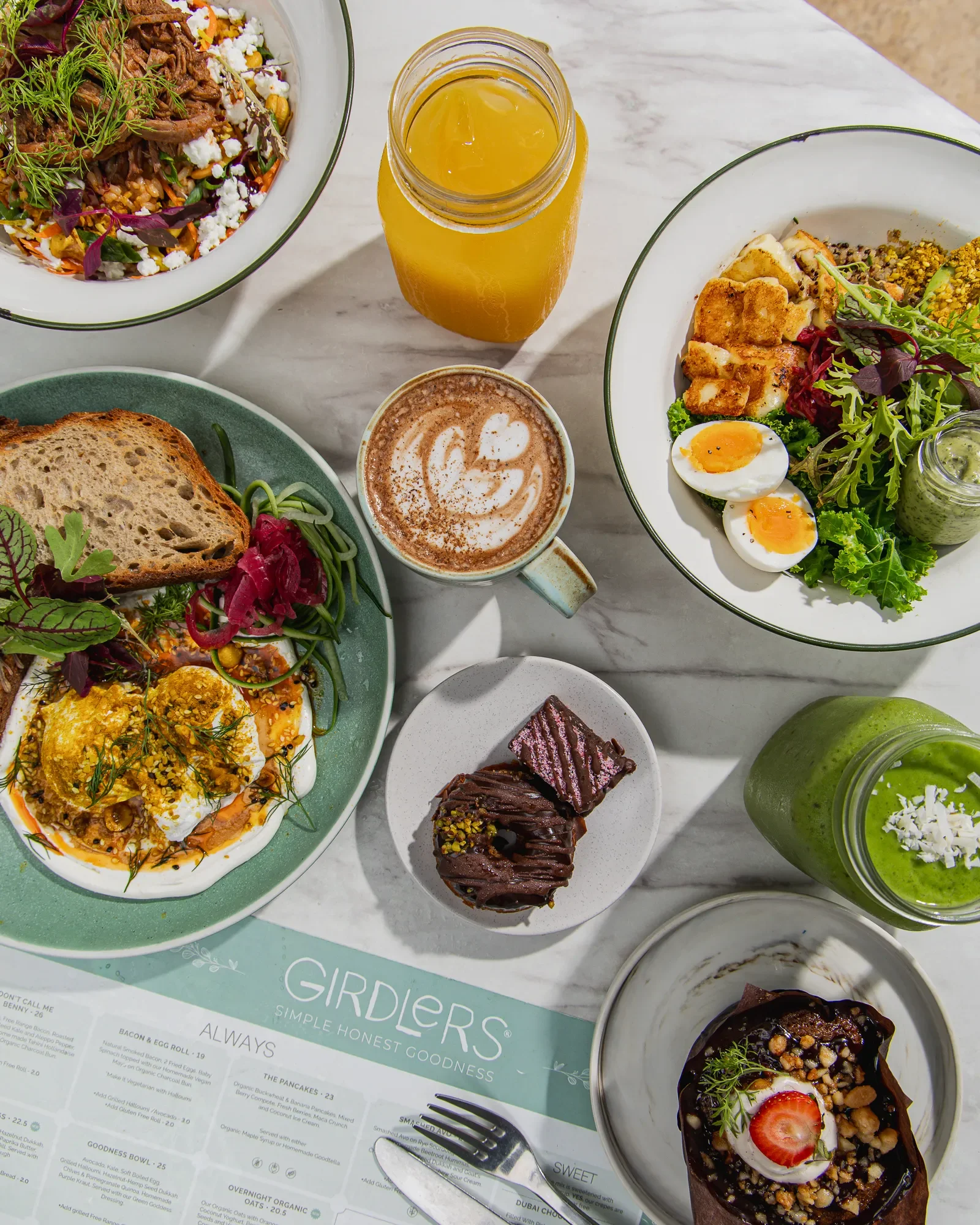 Overhead view of a breakfast or brunch spread featuring bowls of colorful salads, a cup of coffee with foam art, a jar of orange juice, and a green smoothie, with a menu underneath.