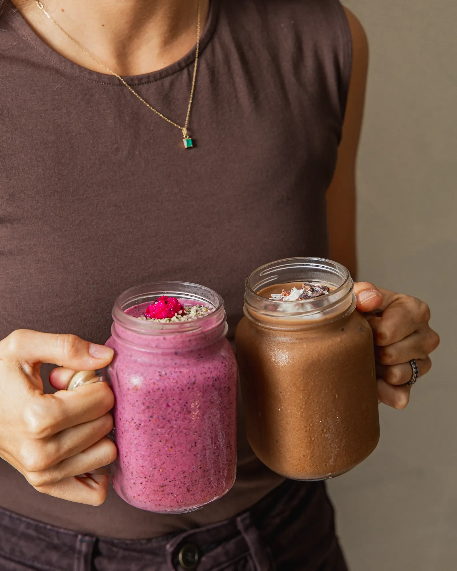 Person in a dark brown sleeveless top holding two mason jars filled with colorful smoothies, one pink with toppings and the other brown, with V-shaped necklace with green pendant visible.