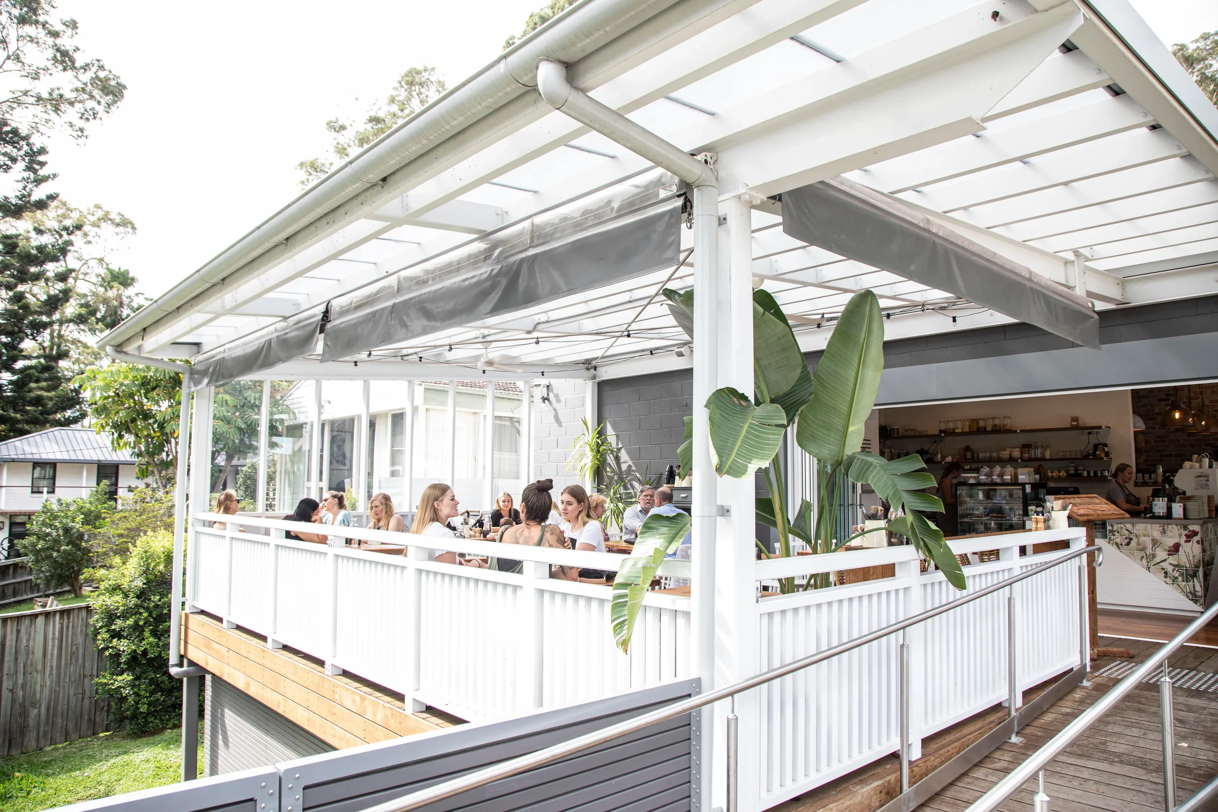 People dining on an outdoor balcony at a restaurant with a white railing and a transparent roof, surrounded by greenery.