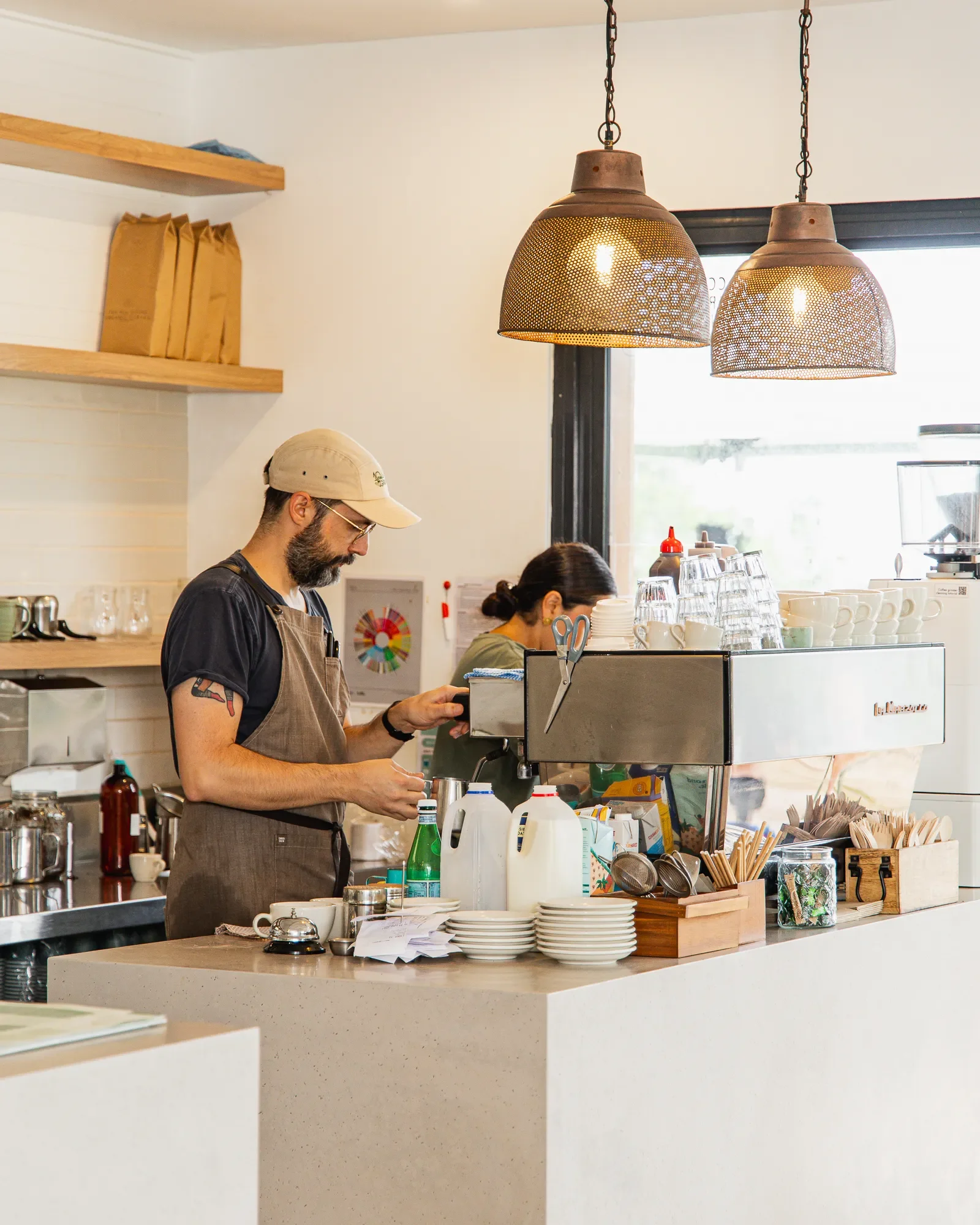 Barista preparing coffee behind a counter in a cafe with shelves, cups, utensils, and hanging lights.