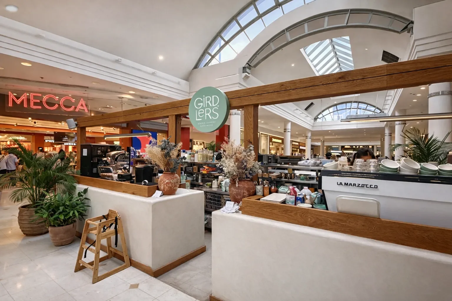 A café booth named 'GIRDLERS' inside a shopping mall, decorated with potted plants and vases, with a sign for 'MECCA' in the background.