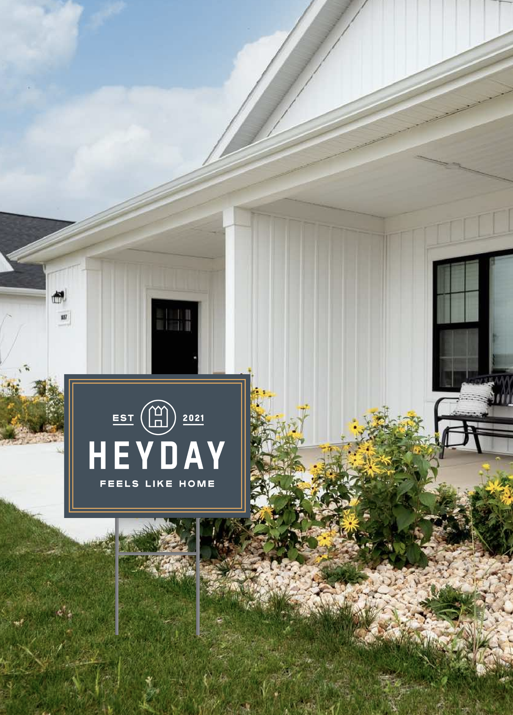 Front yard of a white house with a porch, black window frame, and a logo sign that reads "Heyday, feels like home," surrounded by yellow flowers and green grass.