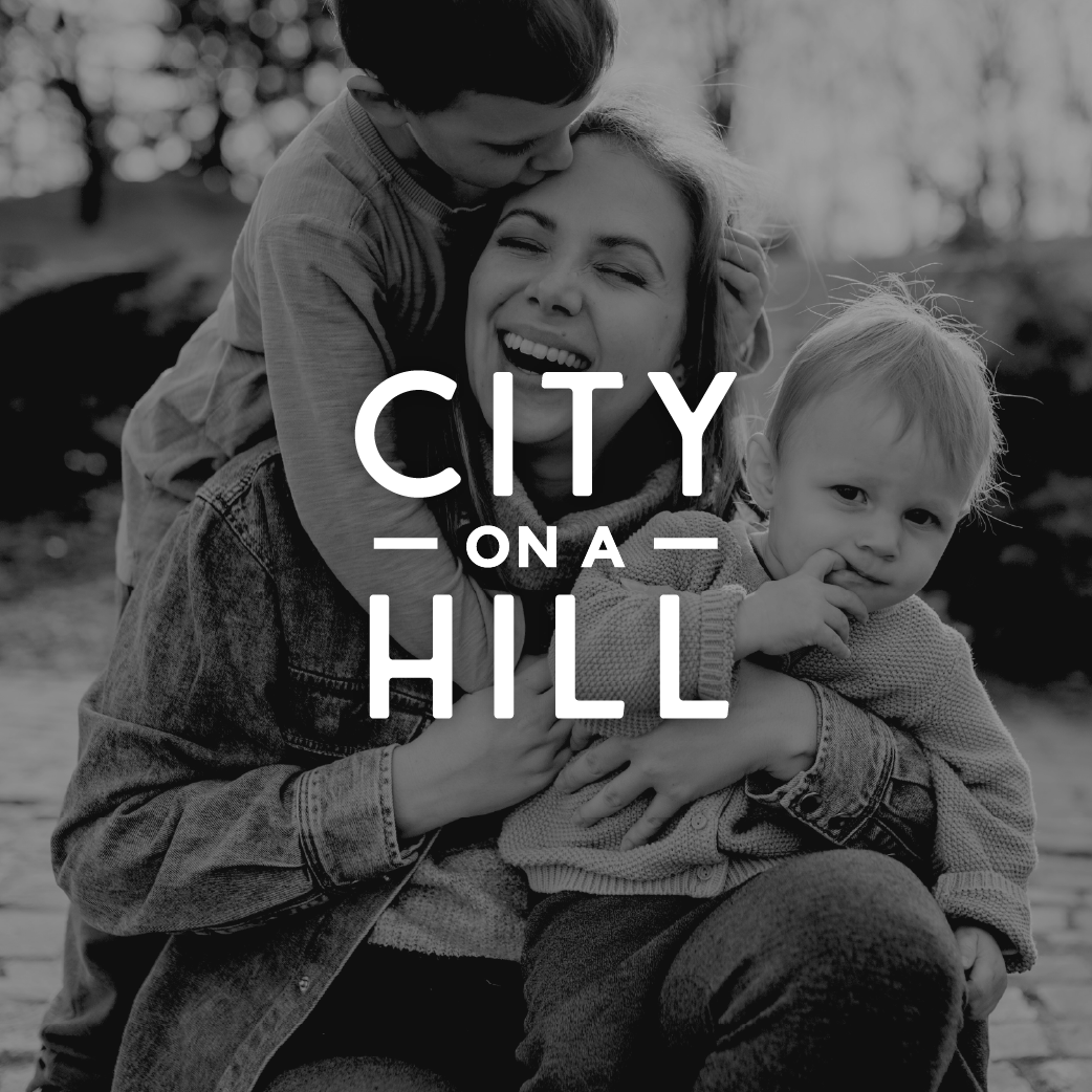 A woman sitting outdoors with two young children, one hugging her from behind and the other sitting on her lap, all smiling and enjoying a moment together.