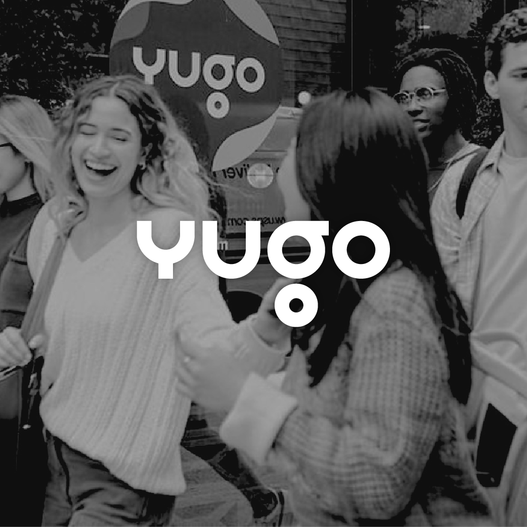 Group of diverse young adults laughing and talking outdoors in front of an 'Yugo' sign.