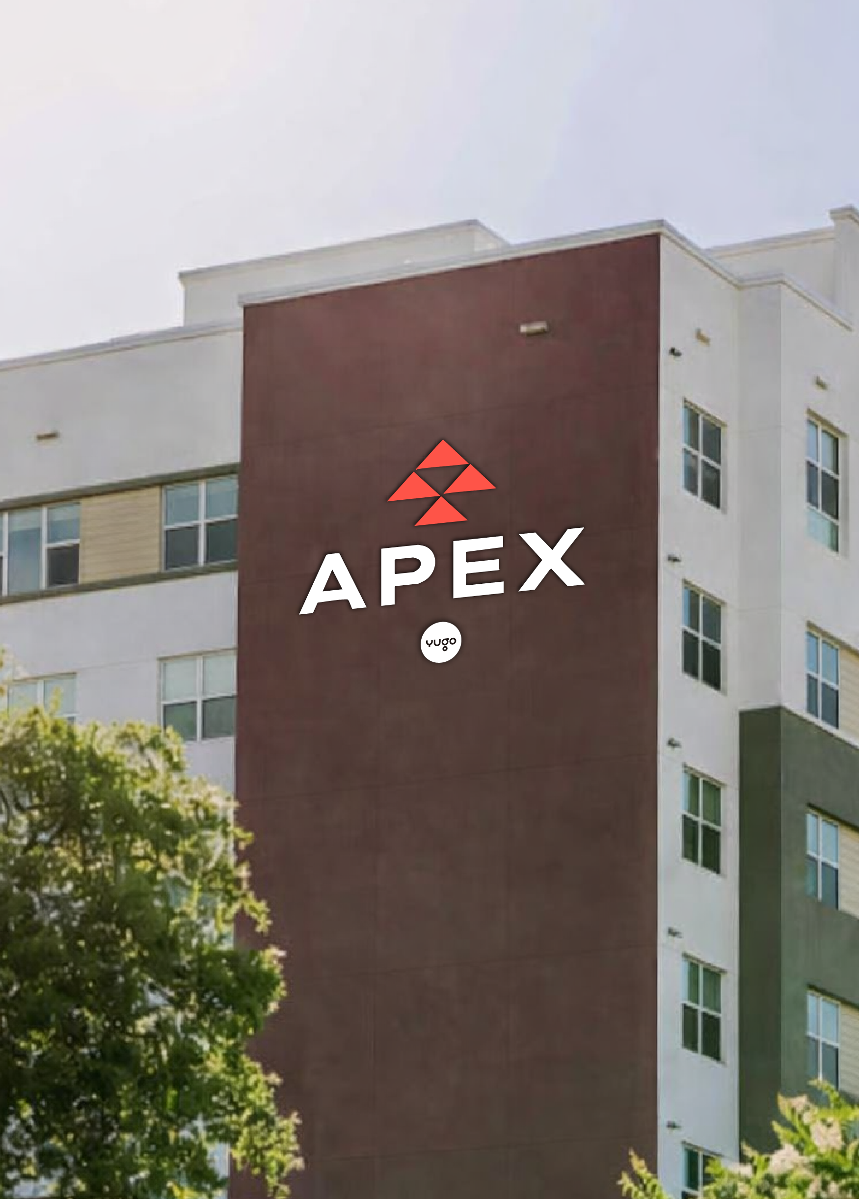 Apartment building with the 'Apex' logo and a stylized red mountain icon on a brown section of the facade, surrounded by other buildings and trees.