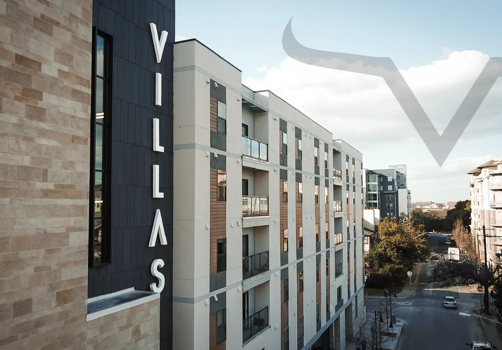 View of a modern apartment building with a sign reading 'Villas' on a black vertical strip, and a nearby street with cars and trees.
