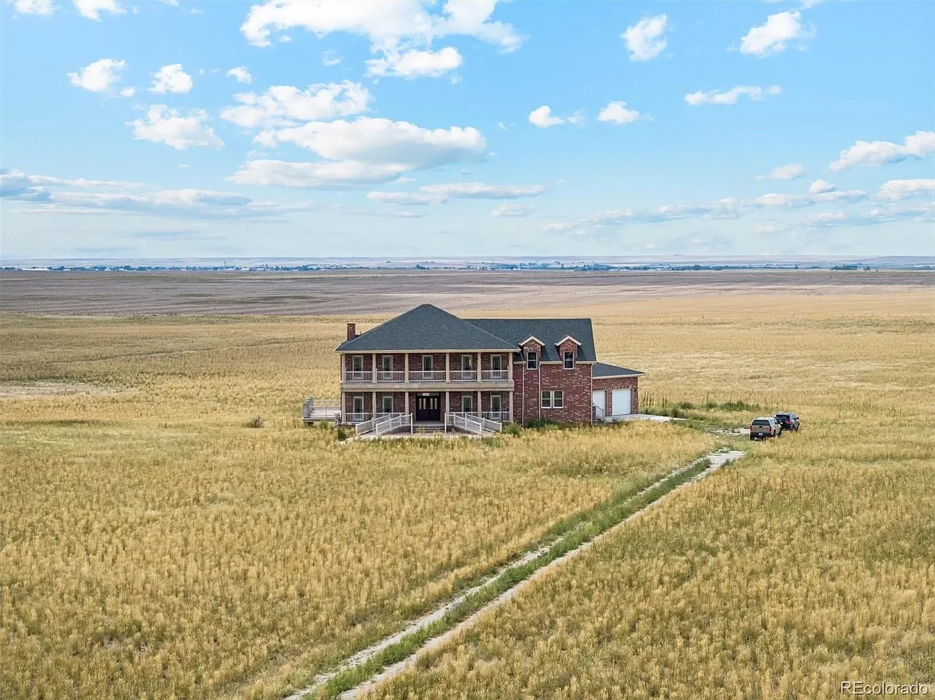 A large two-story brick house with a green roof and a spacious balcony, situated in a vast open field of tall yellow grass under a partly cloudy sky.