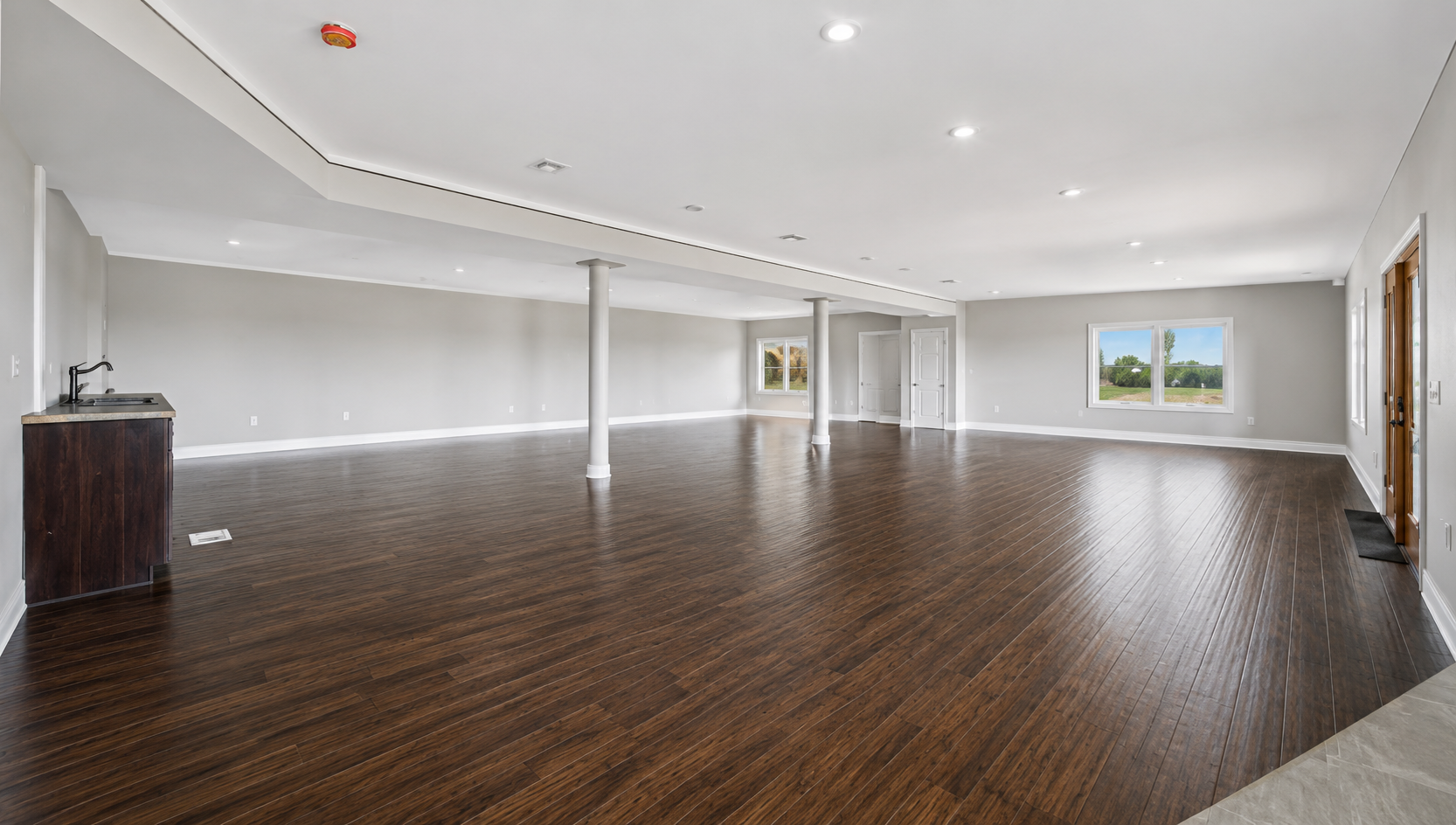 Empty living room with dark hardwood floors, white walls, multiple windows, ceiling lights, and four columns, with a small kitchen area in the corner.