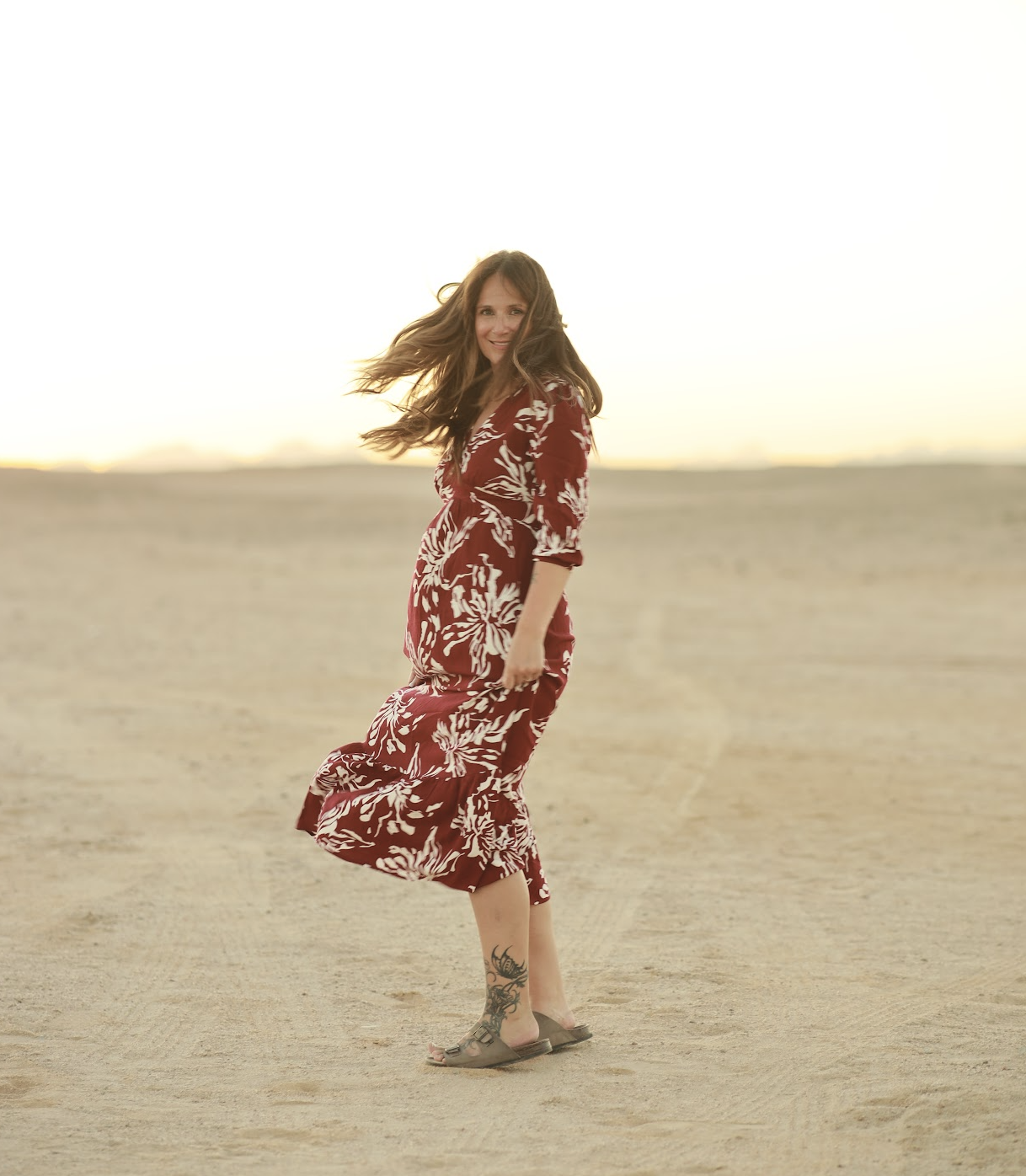 Woman with long brown hair wearing a red and white patterned dress, standing on a sandy beach with a sunset in the background, smiling and twirling.
