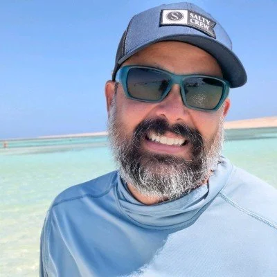 Close-up of a man with a beard wearing sunglasses, a blue cap with a logo, and a blue shirt, smiling at the beach with clear water and blue sky in the background.