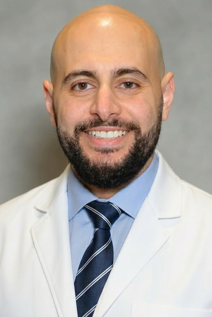 A smiling man with a beard and no hair, wearing a white coat, light blue shirt, and dark striped tie, standing against a gray background.