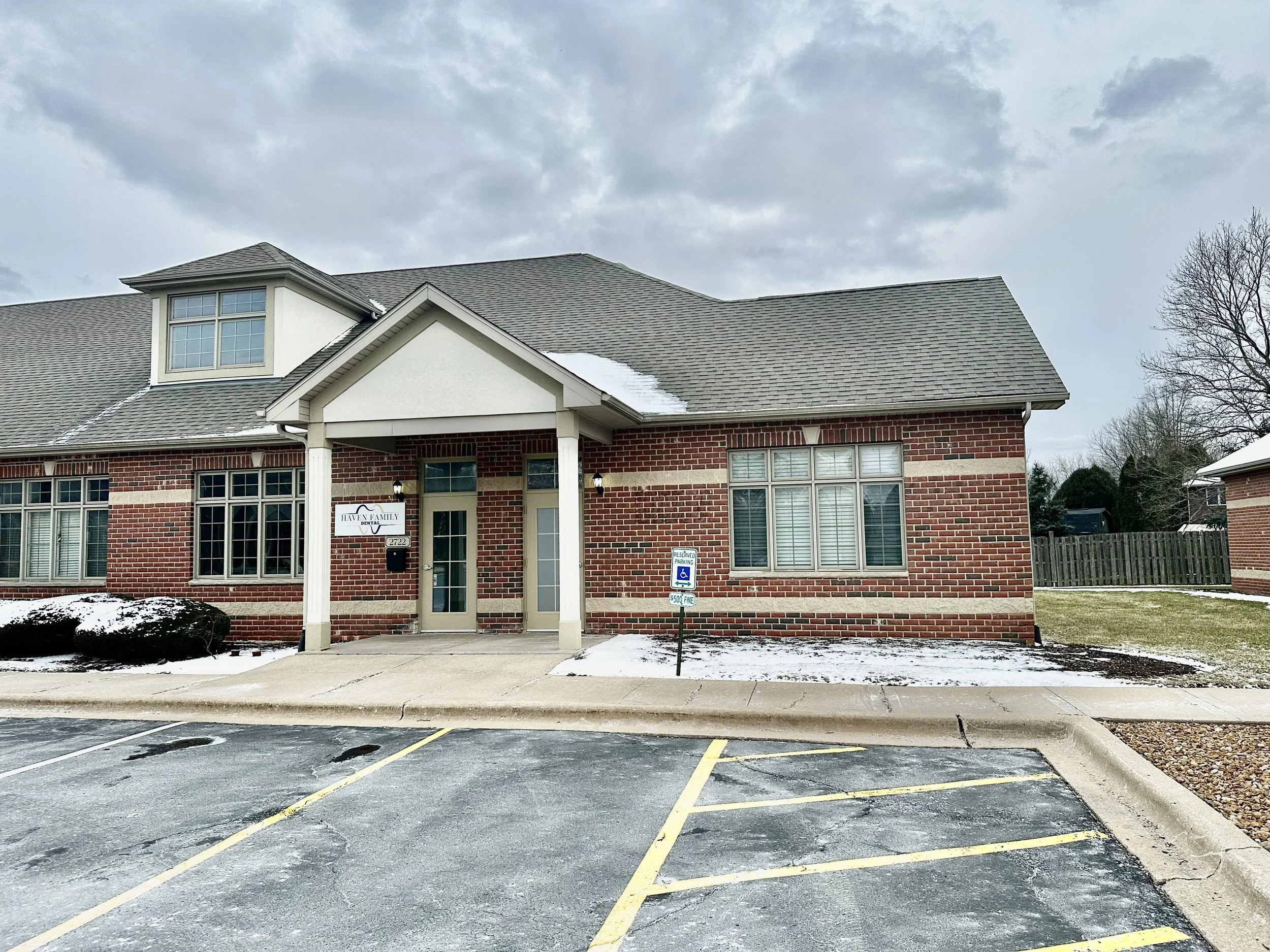 Two-story brick building with a parking lot in front, a handicapped parking sign, snow on the ground, and an overcast sky.