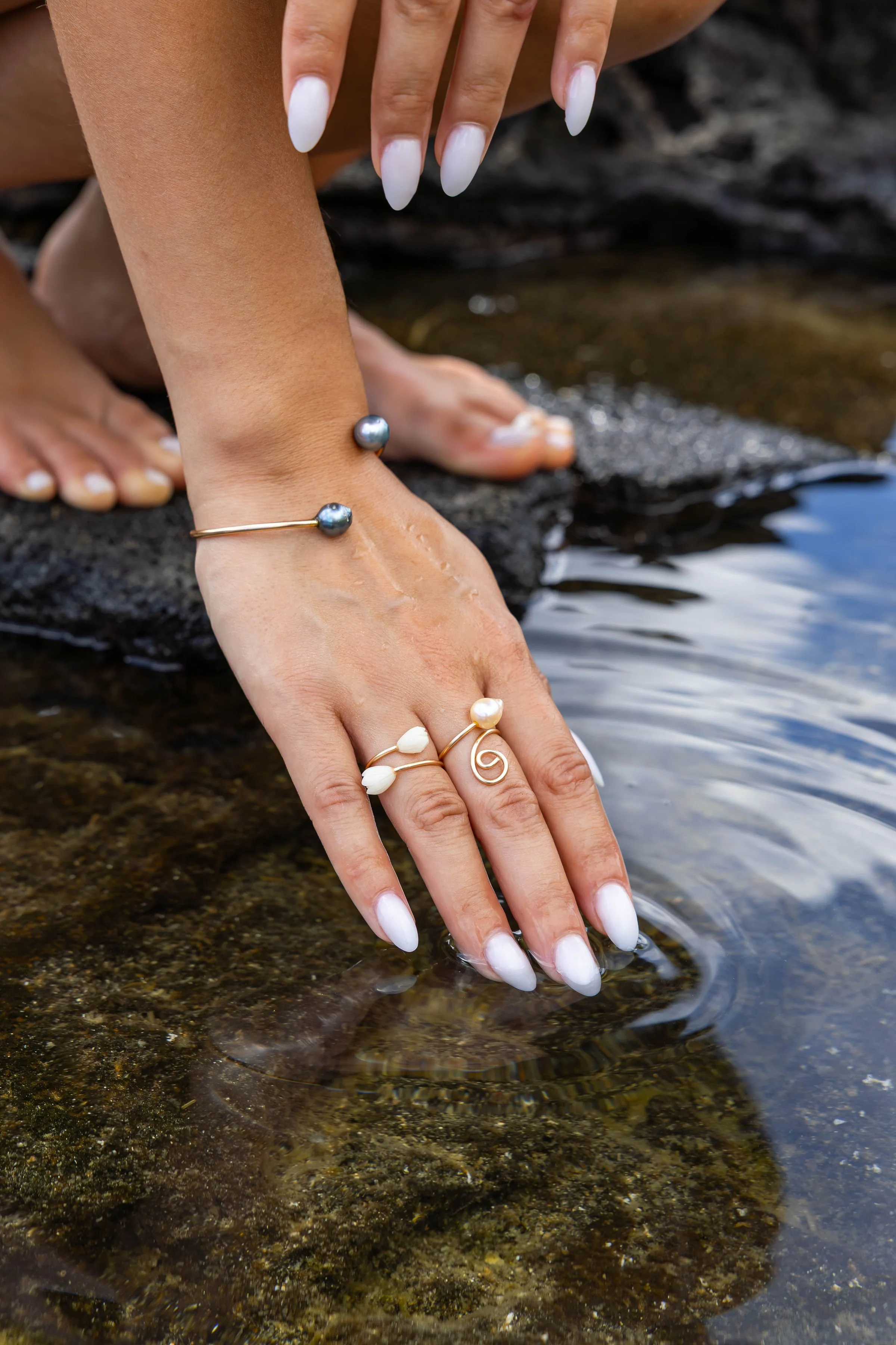 Close-up of a woman's hand and wrist with jewelry, resting on wet rocks near a shallow stream or river. The jewelry includes pearl and gold rings, and a pearl bracelet.