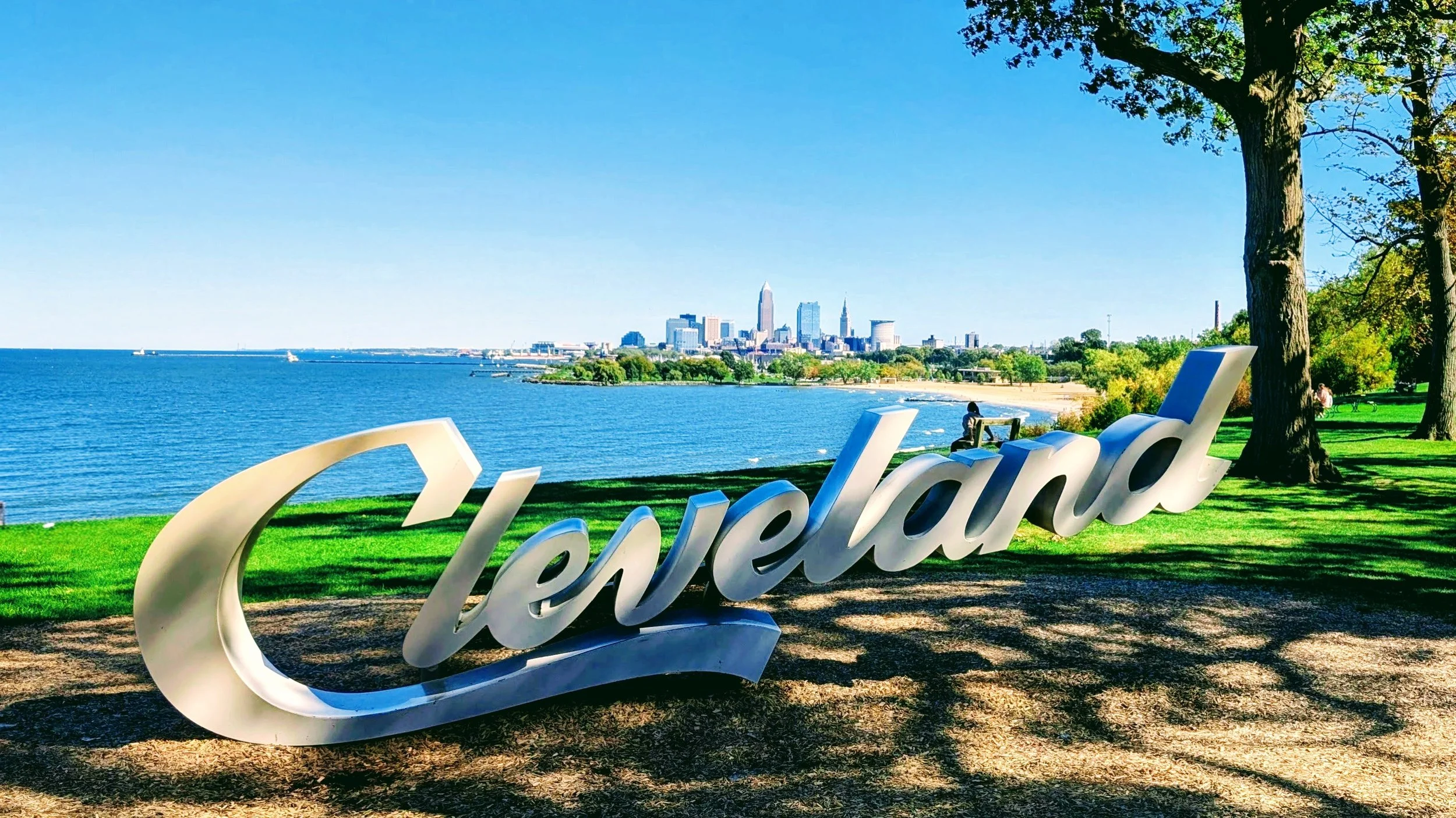 Large decorative sign spelling 'Cleveland' in a park by Lake Erie, with the Cleveland skyline in the background and trees and benches nearby.