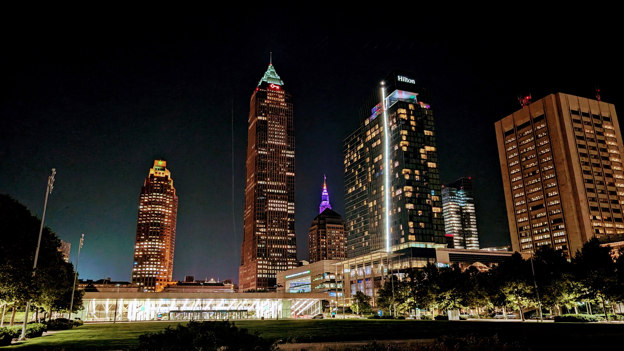 Nighttime view of downtown Atlanta city skyline with illuminated skyscrapers, including the Bank of America Plaza with its green spire, and the Hilton hotel with its logo, as well as other tall buildings and trees in the foreground.