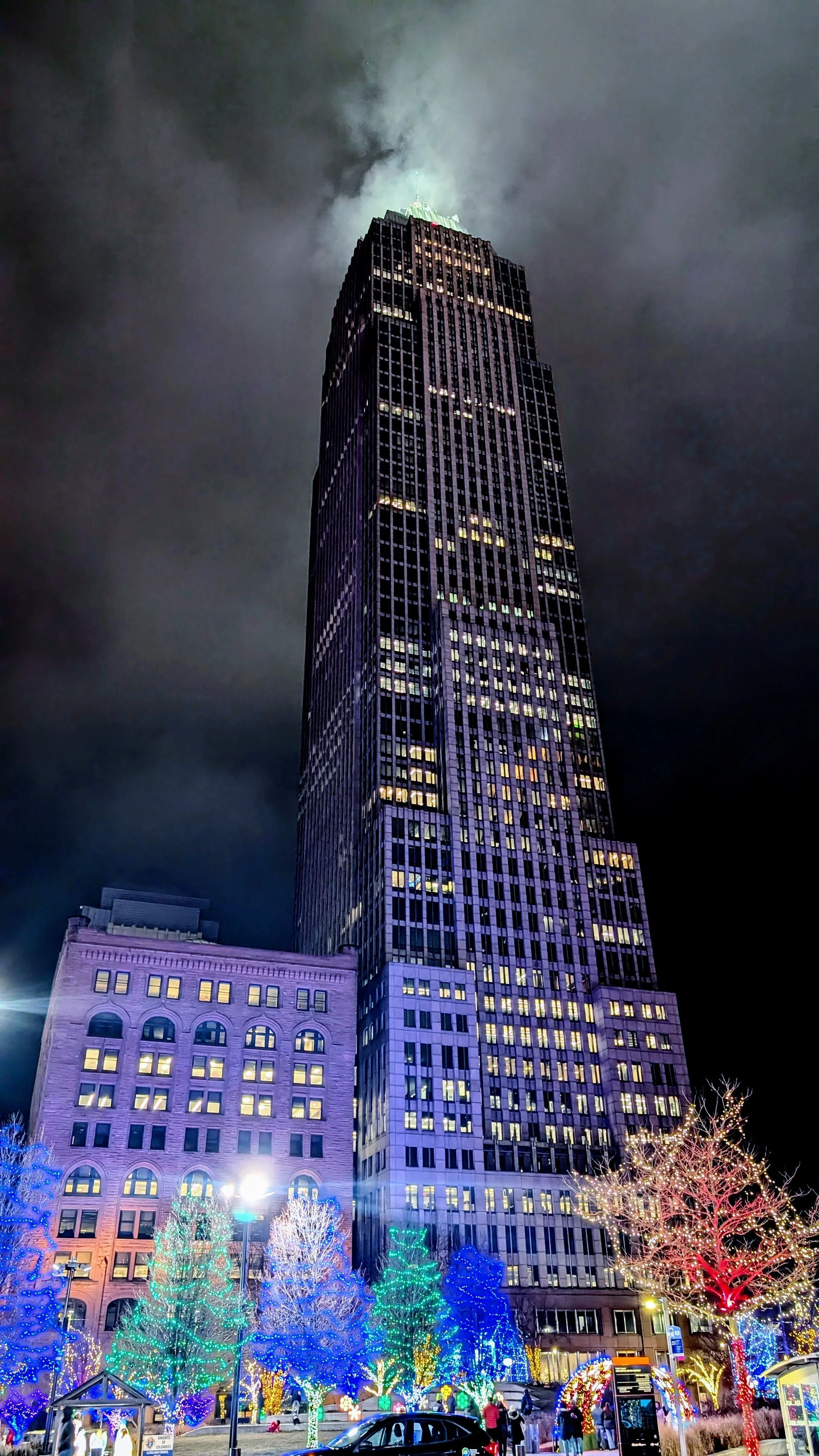 Night view of a tall skyscraper with illuminated windows and colorful holiday lights on nearby trees in the foreground.