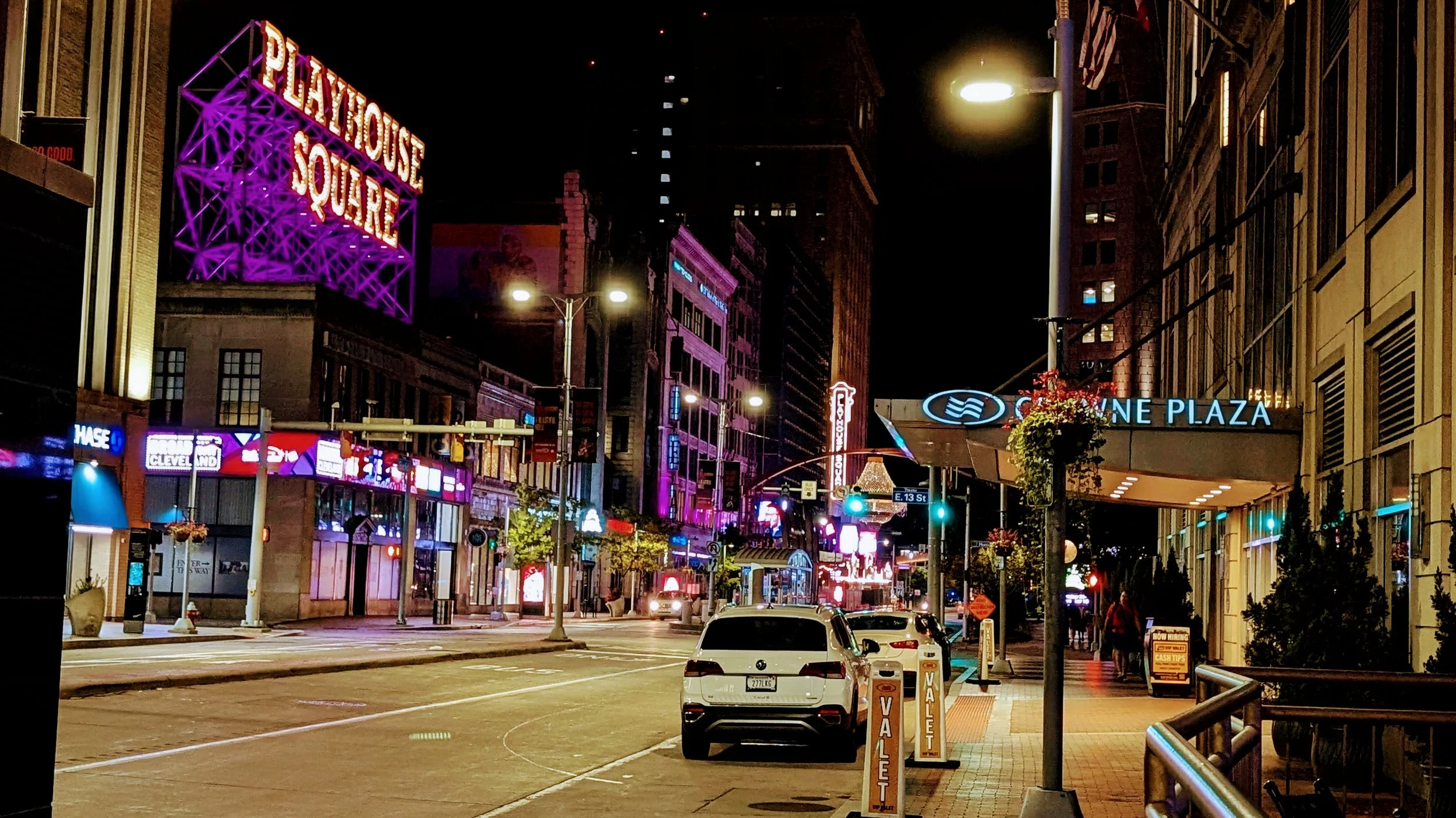 Nighttime street scene with illuminated signs and buildings, including a large purple neon sign reading 'Playhouse Square' and a blue sign for 'Cleveland Plaza,' with a few cars parked on the street and some pedestrians on the sidewalk.