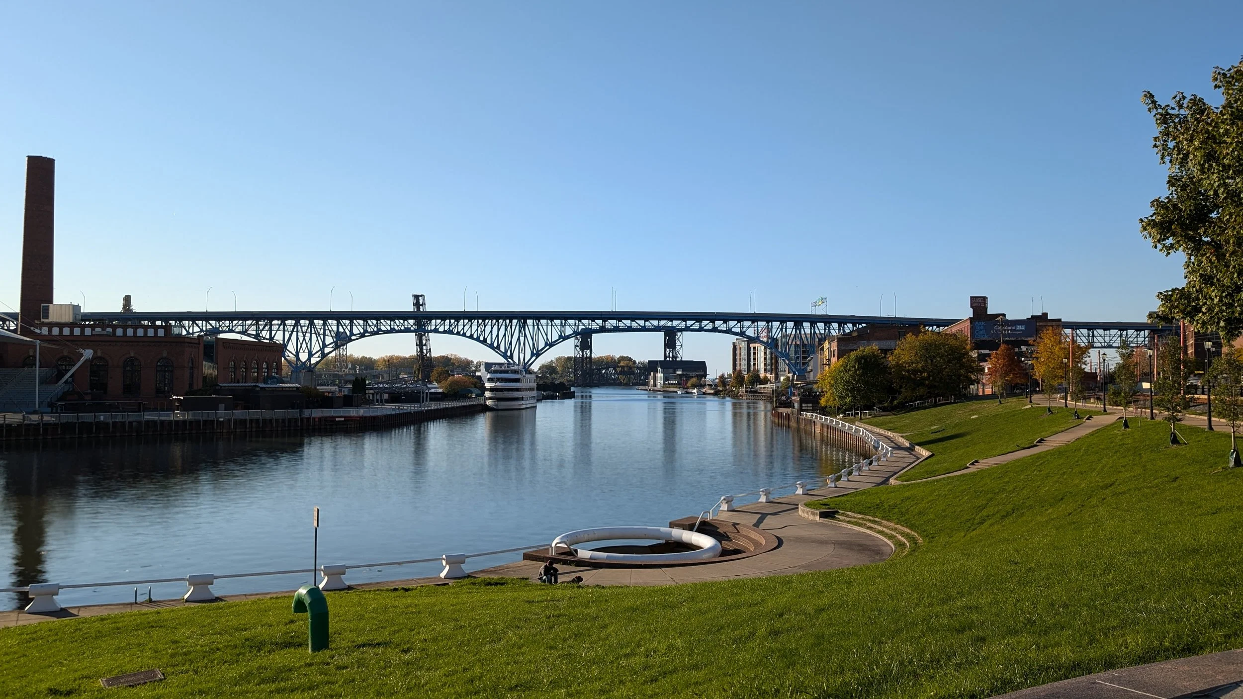 A riverside cityscape with a bridge over the river, boats docked along the side, and a park with grassy hills and trees.