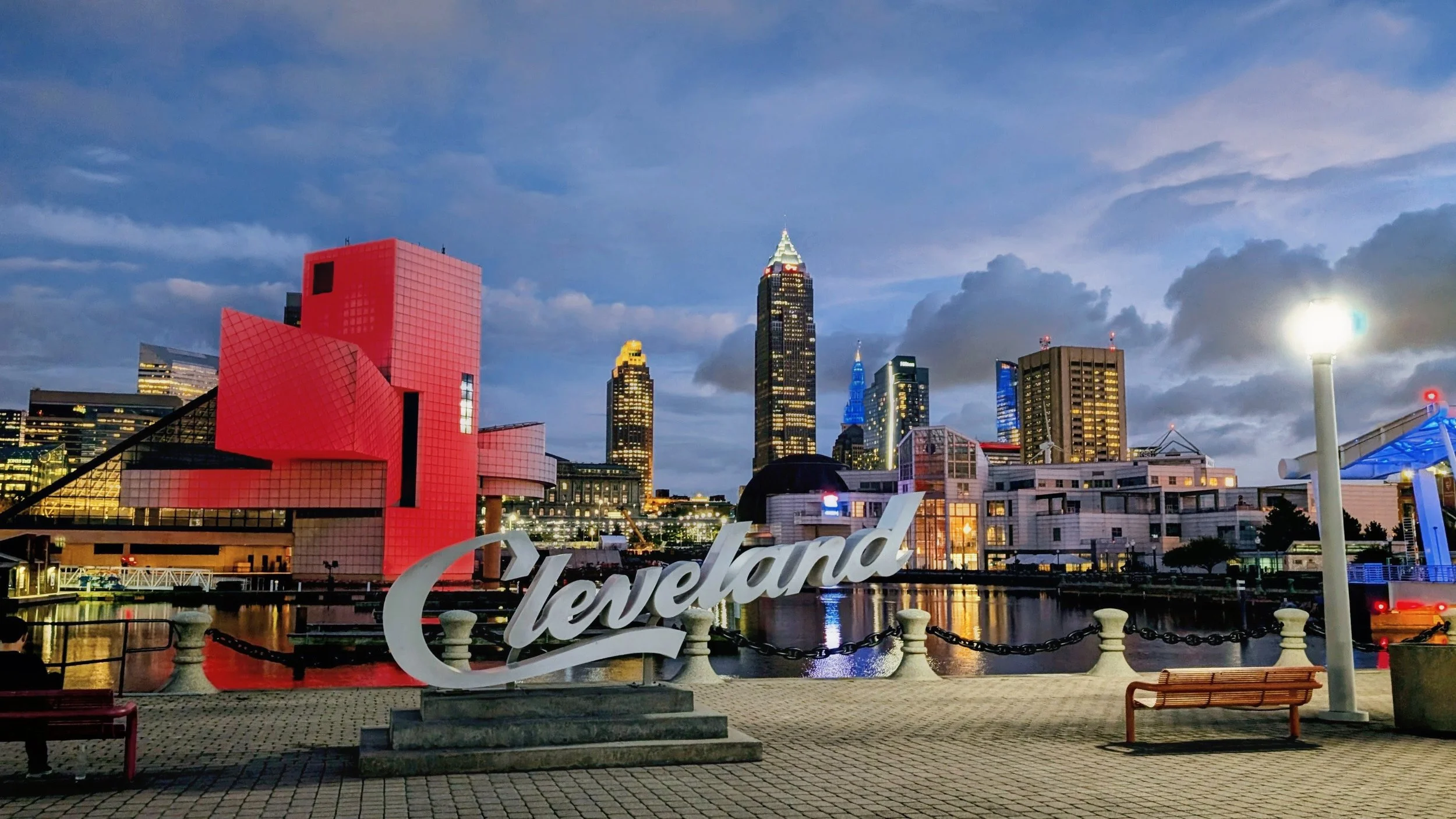 Cleveland city skyline at dusk featuring the Rock and Roll Hall of Fame, with downtown skyscrapers in the background and a waterfront promenade in the foreground.