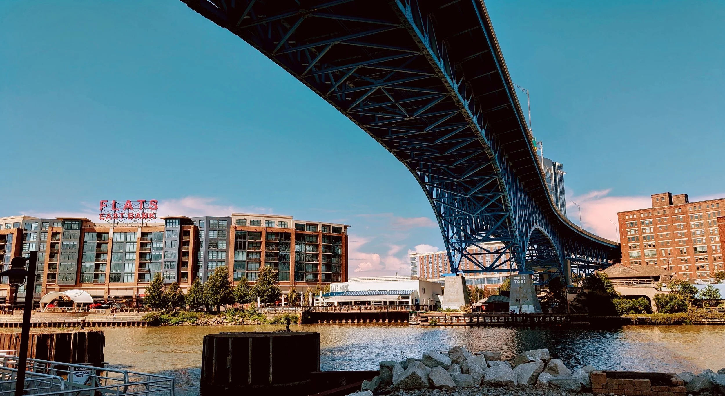 View of a cityscape with a river, a large blue steel arch bridge, residential buildings, and a sign that reads 'FLATS EAST BANK' in the background. The sky is clear with a few clouds.