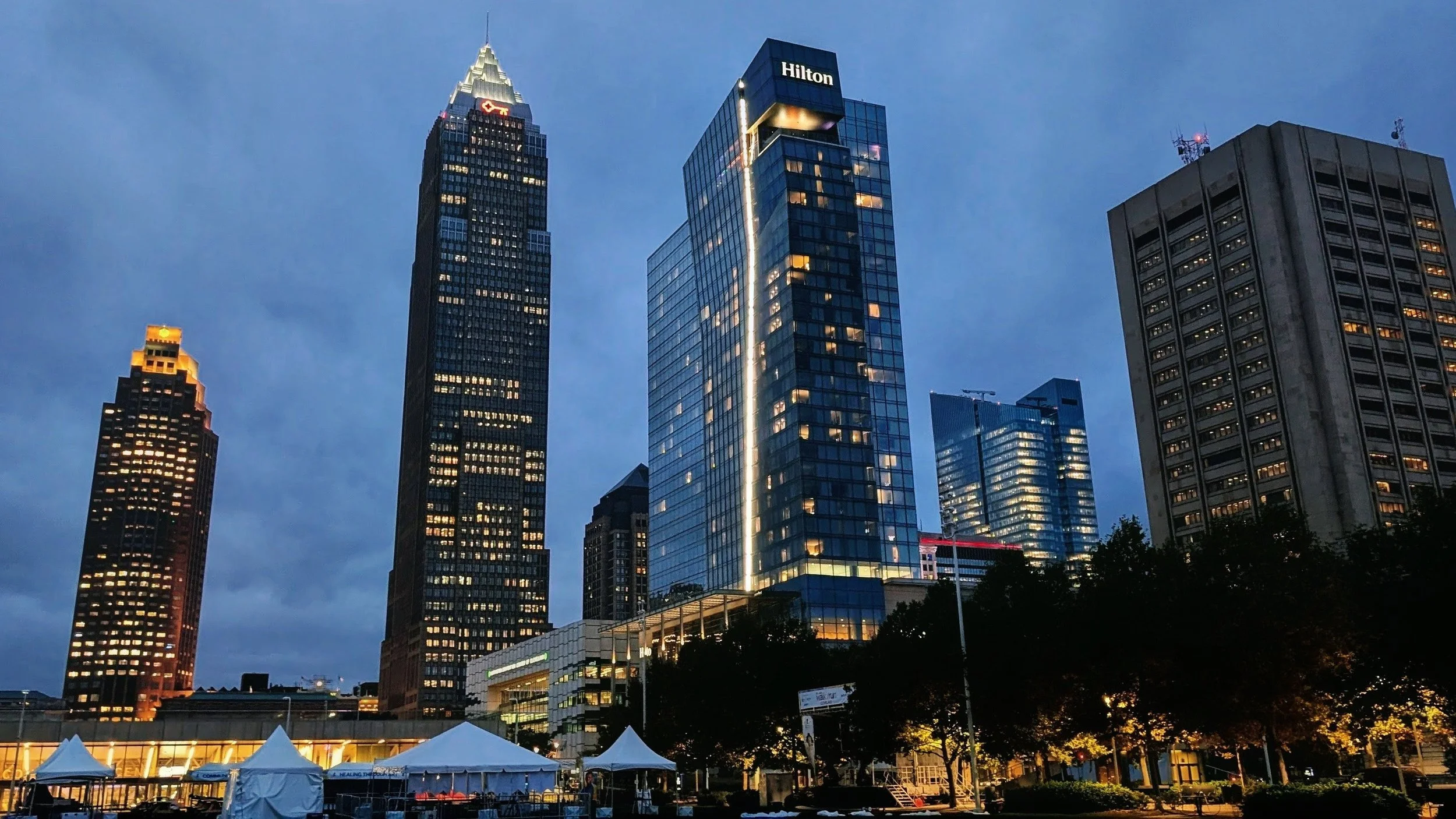 City skyline at dusk with illuminated skyscrapers and trees in the foreground.