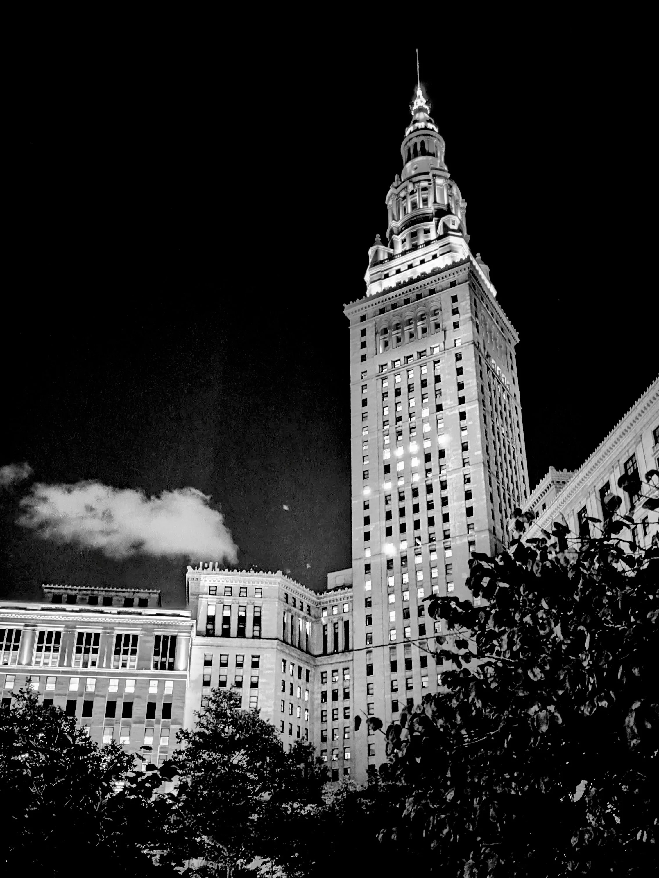 Night black-and-white photo of the Empire State Building in New York City, illuminated against the dark sky with some trees in the foreground.